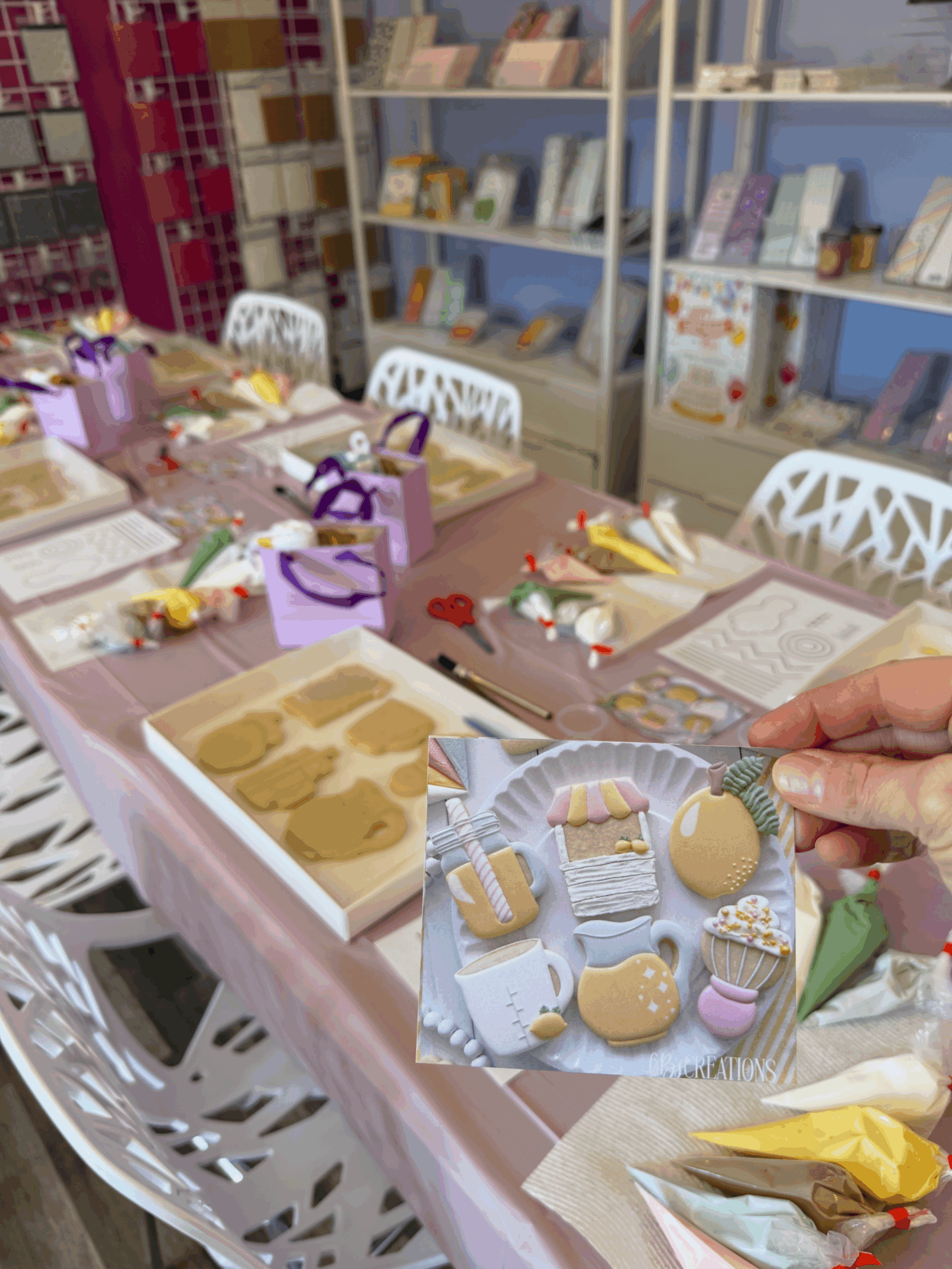 Person decorating cupcakes with colorful toppings on a table with gift boxes and baking supplies, shelves in background.