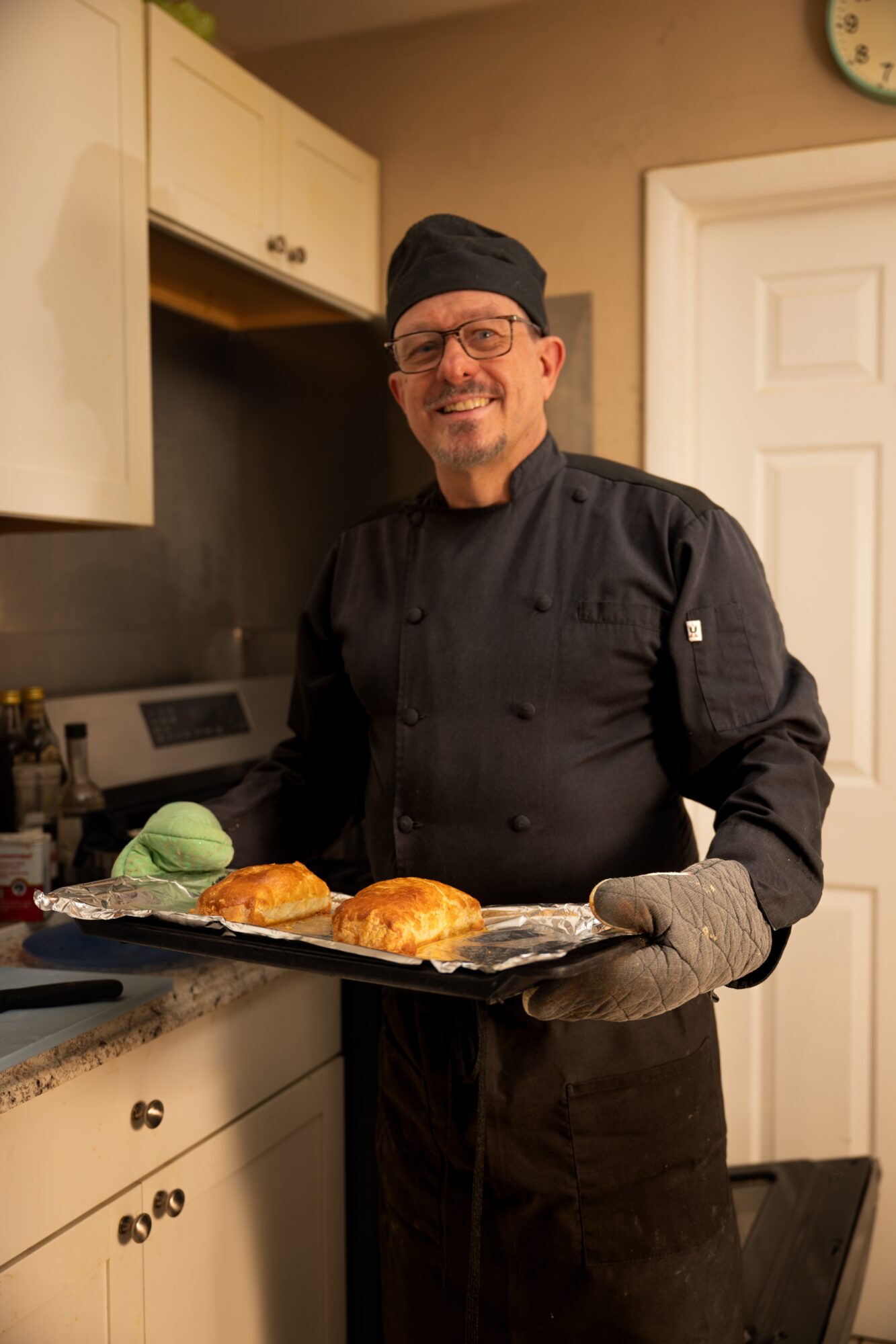 Chef in black uniform and hat holding a tray with bread and green cloth, smiling in a kitchen.