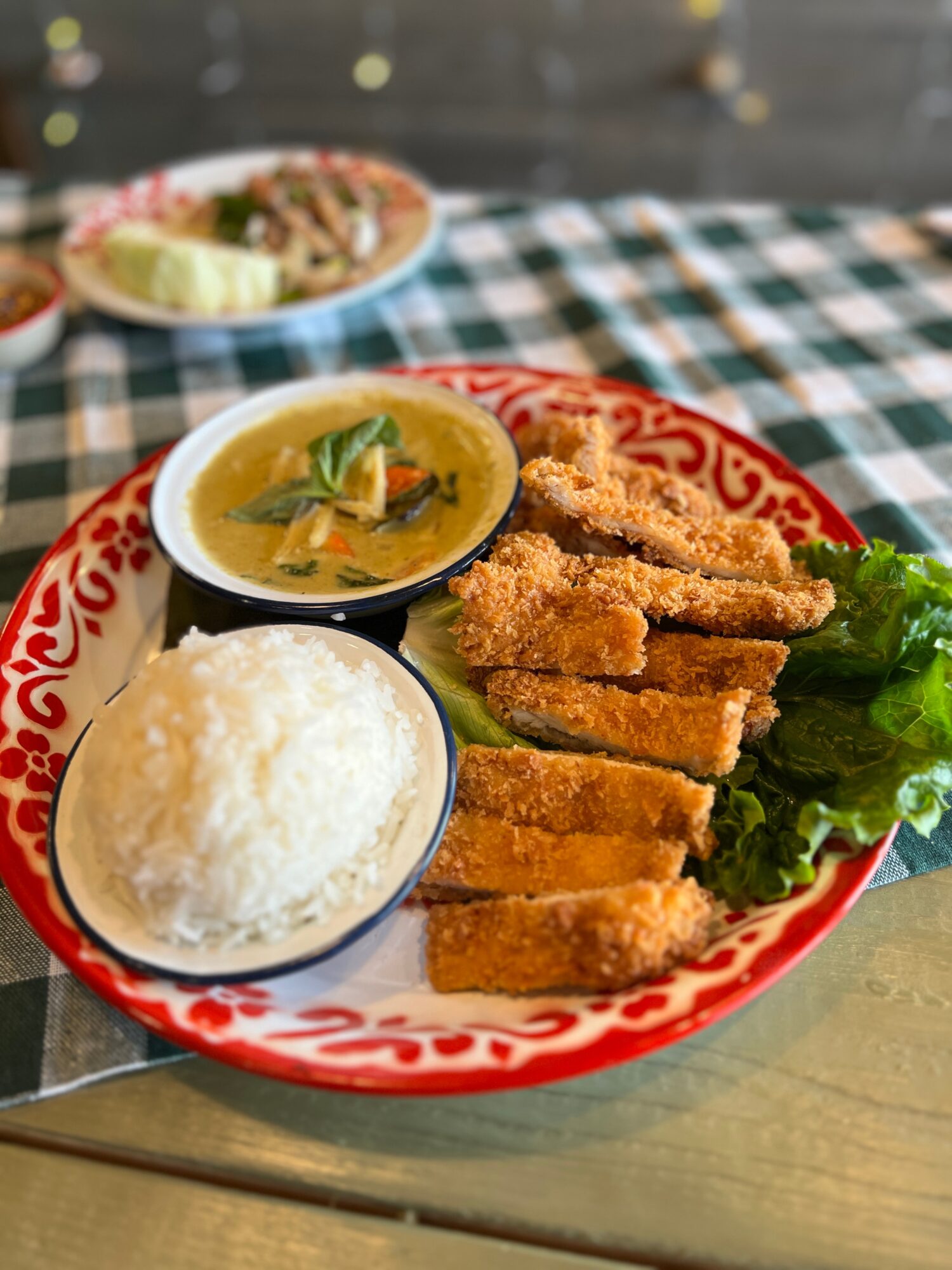 Plate with fried fish, rice, yellow curry, and lettuce on a red and white patterned dish, on a checkered tablecloth.