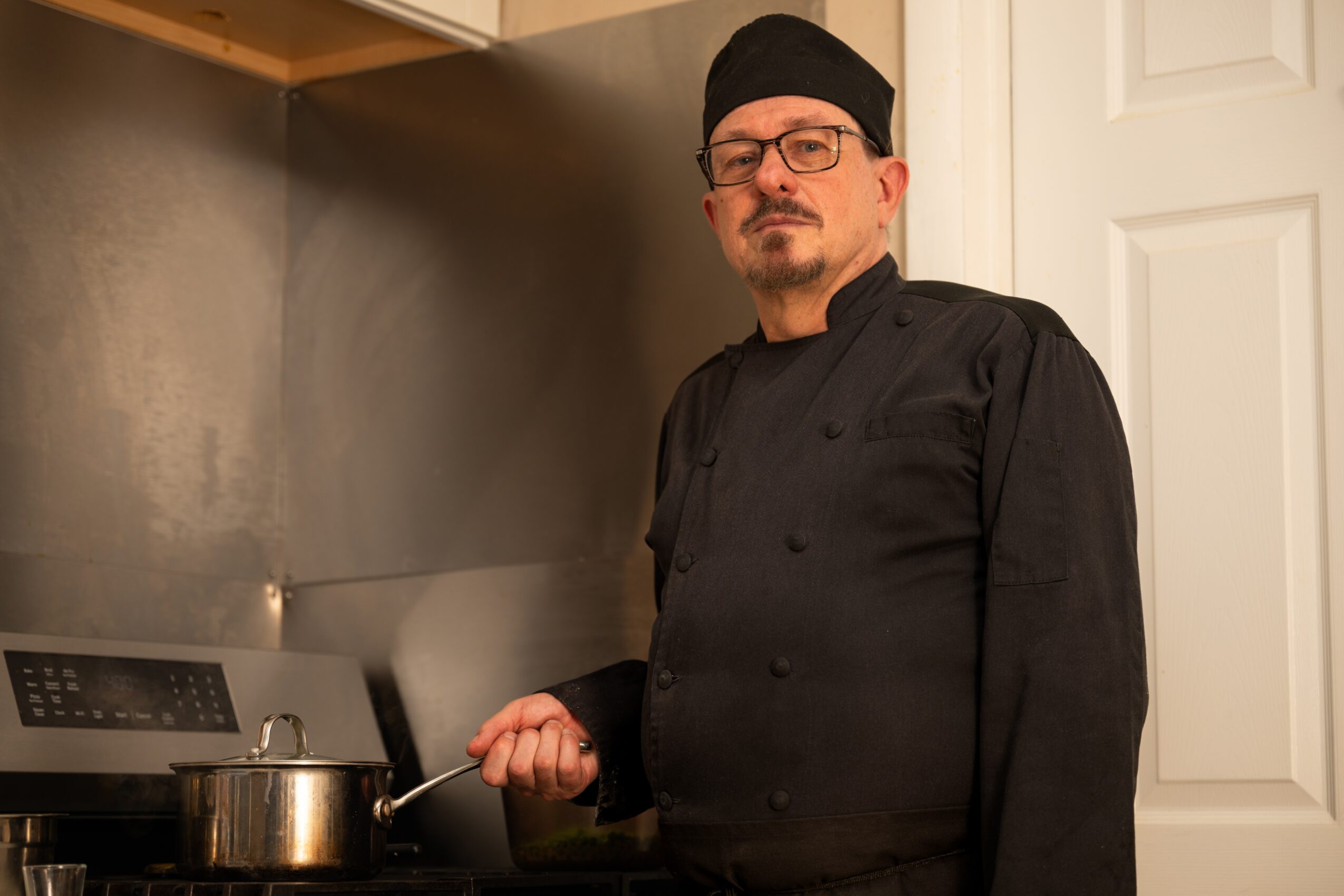 Chef in black uniform and hat holding a spoon in a kitchen, standing near a stove and a white door.