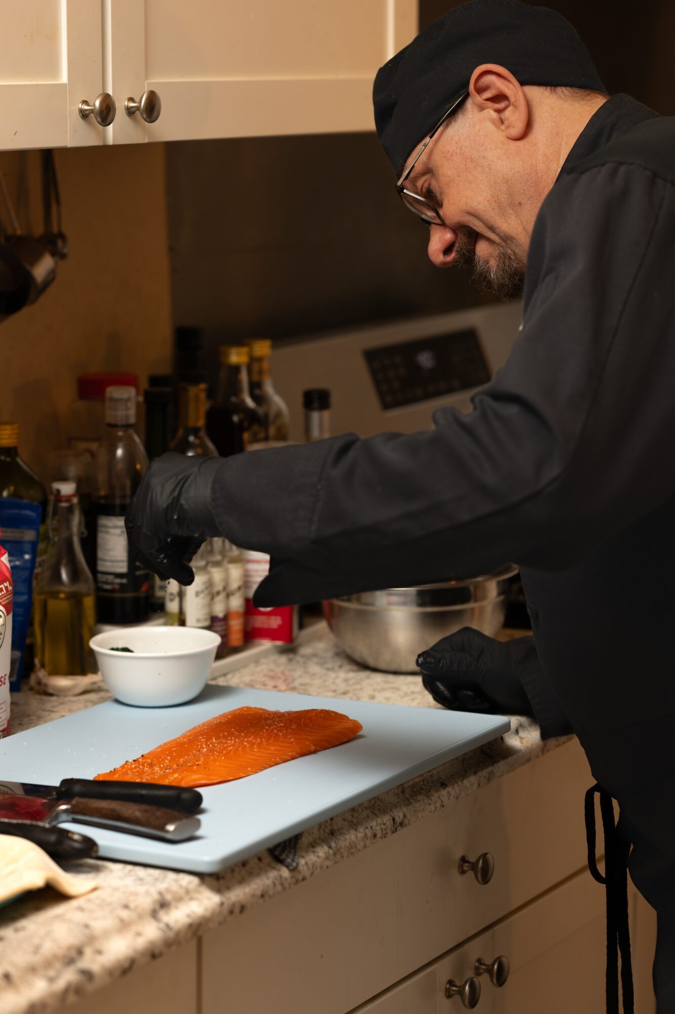 Chef in black uniform and glasses preparing food in a kitchen with various bottles and ingredients, focusing on a cutting board with fish.