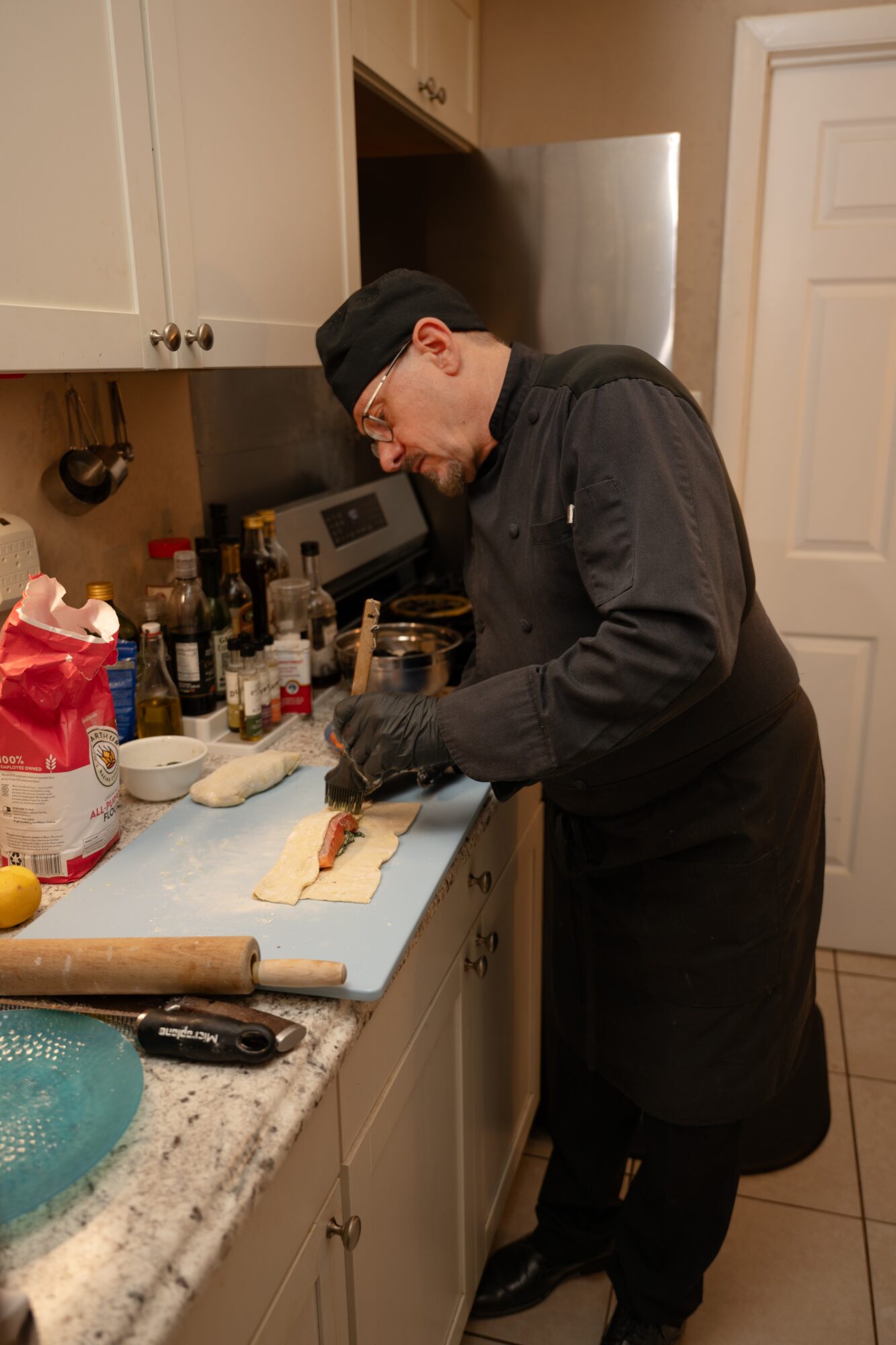 Person wearing black chef's coat and hat preparing food on a kitchen counter with various ingredients and utensils.