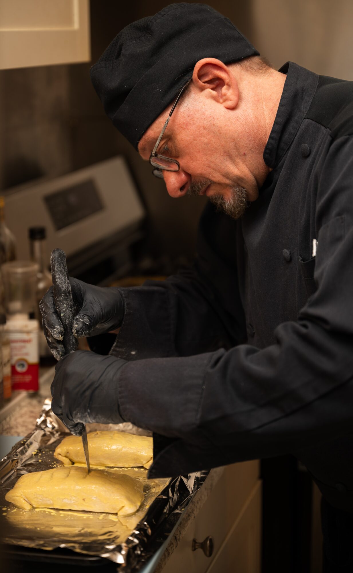Chef wearing black uniform and hat preparing food in a kitchen, focused on his task, with kitchen appliances in background.
