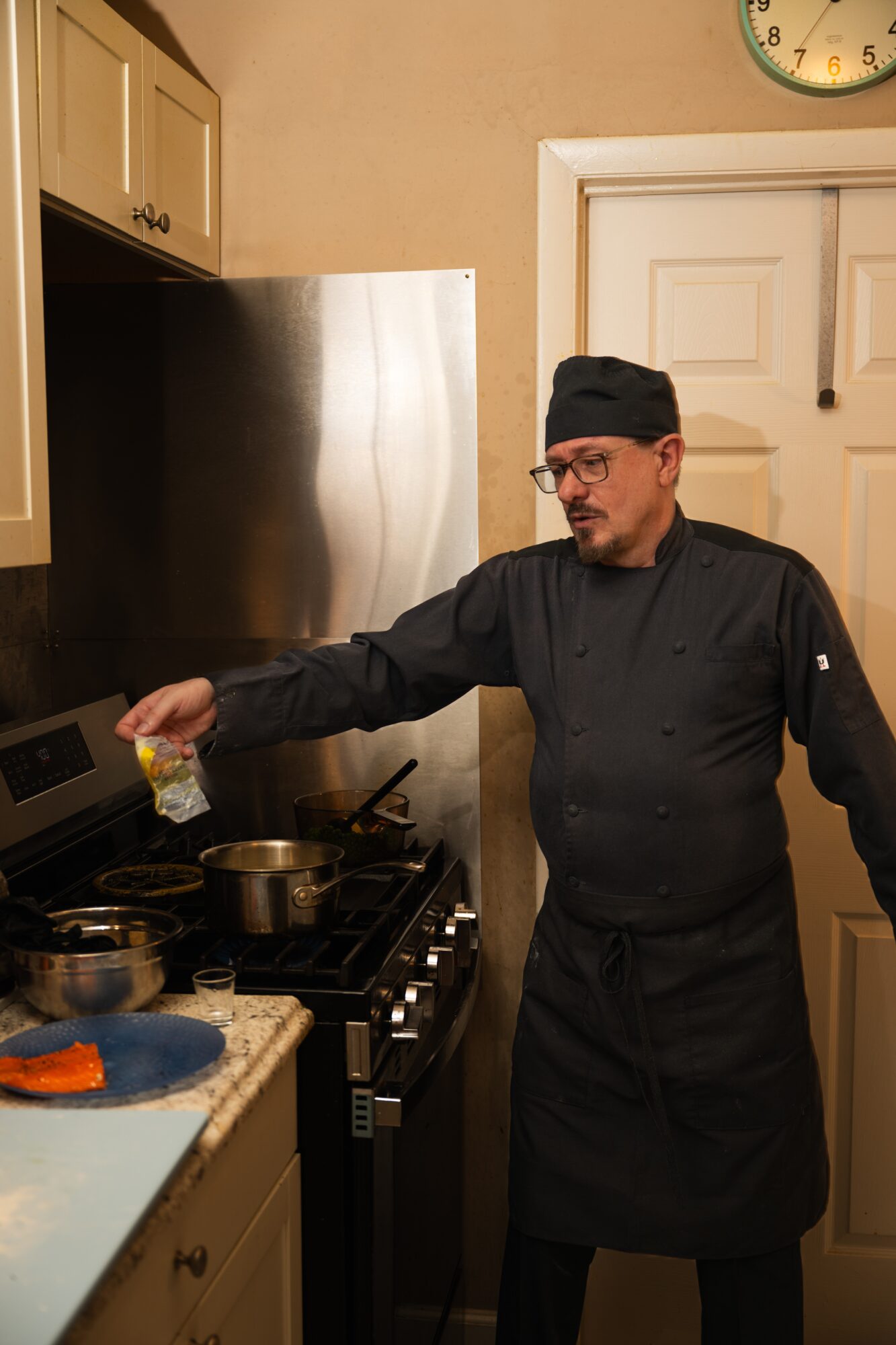 Chef in black uniform and hat cooking in a kitchen with stainless steel appliances and beige walls.