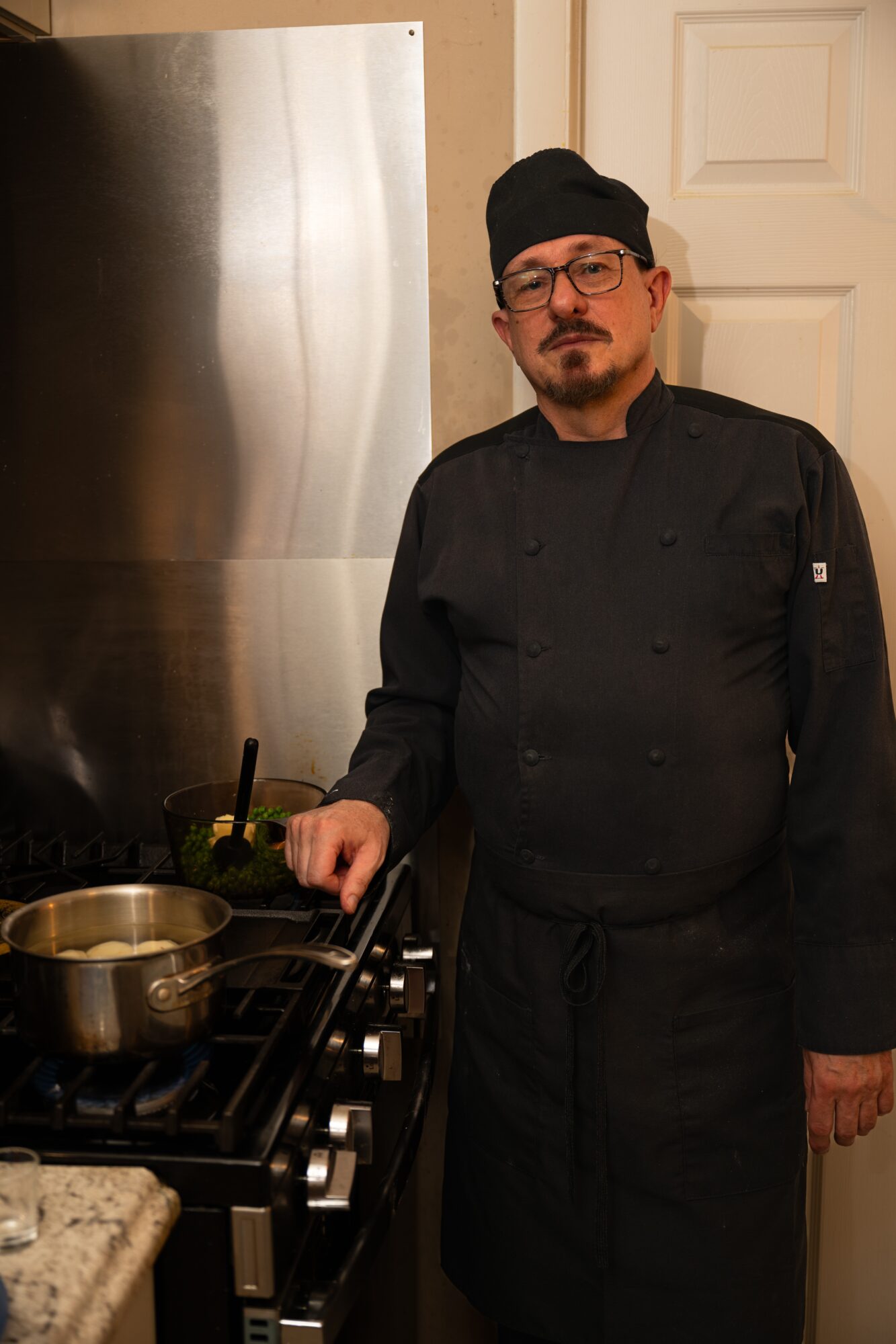 Chef in black uniform and hat standing next to stove in kitchen, looking at camera.