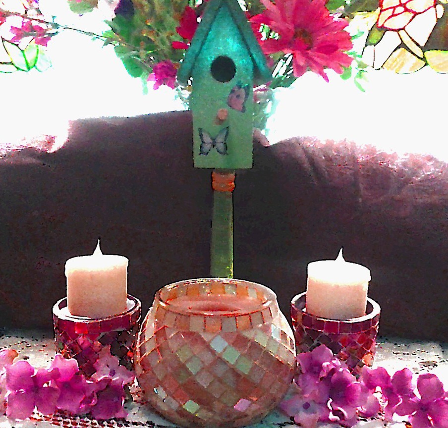 Decorative birdhouse with flowers, candles, and a mosaic bowl on a table, with a stained glass window in the background.