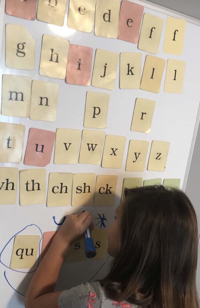 Child writing on a whiteboard with alphabet and phonics cards, some cards are red, yellow, and blue, with letters and sounds.