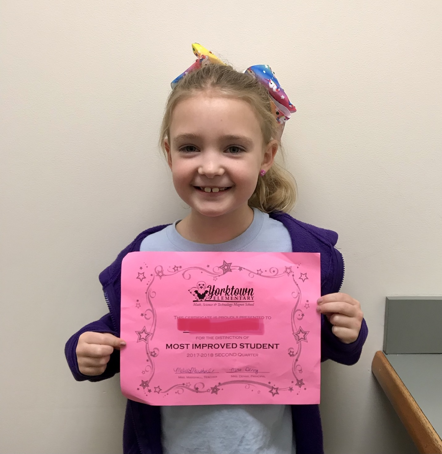 Young girl holding a pink certificate, smiling, with a colorful bow in her hair, standing against a plain wall.