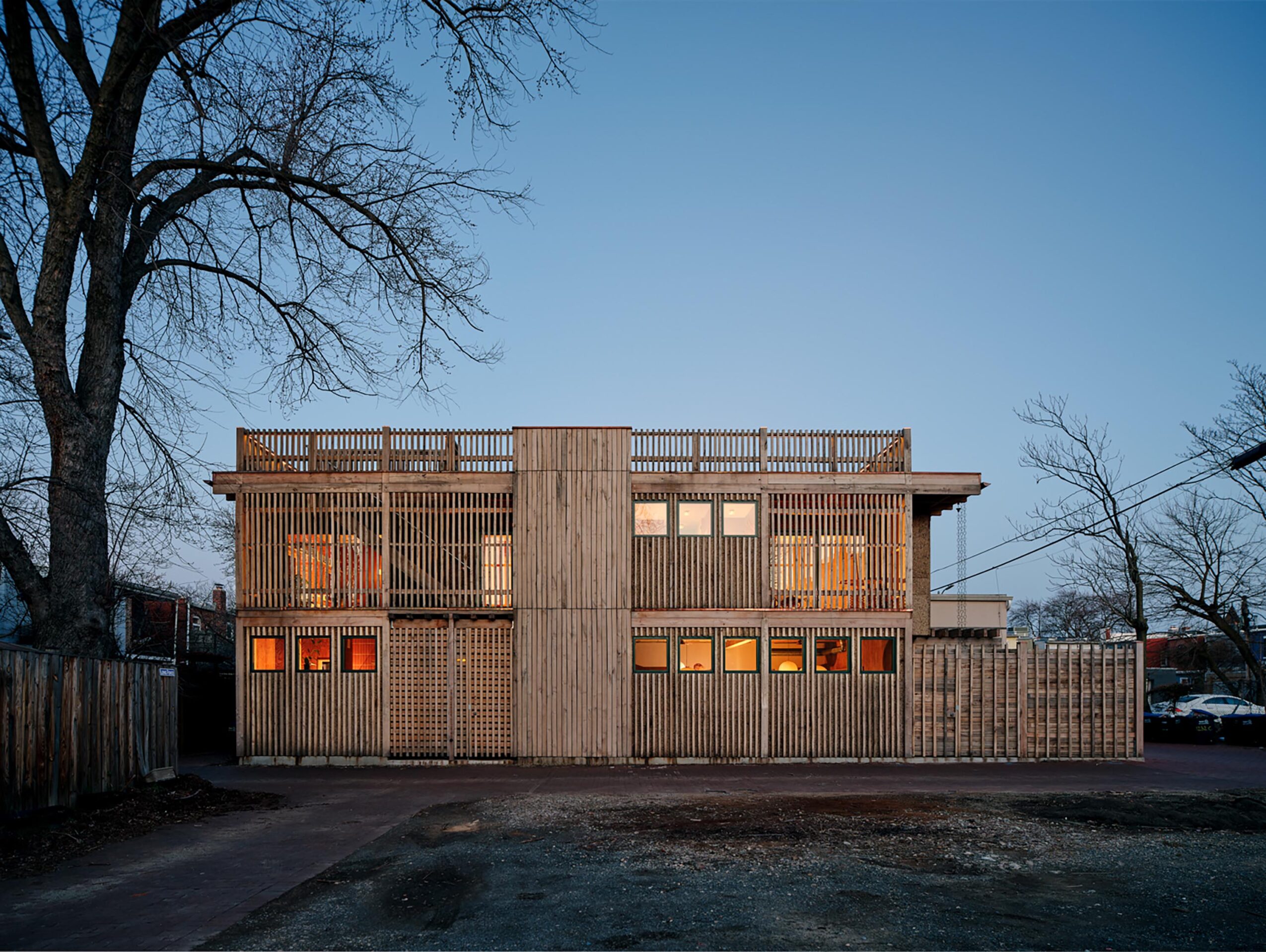 Two-story wooden building with a fence, trees on sides, and a clear sky background.