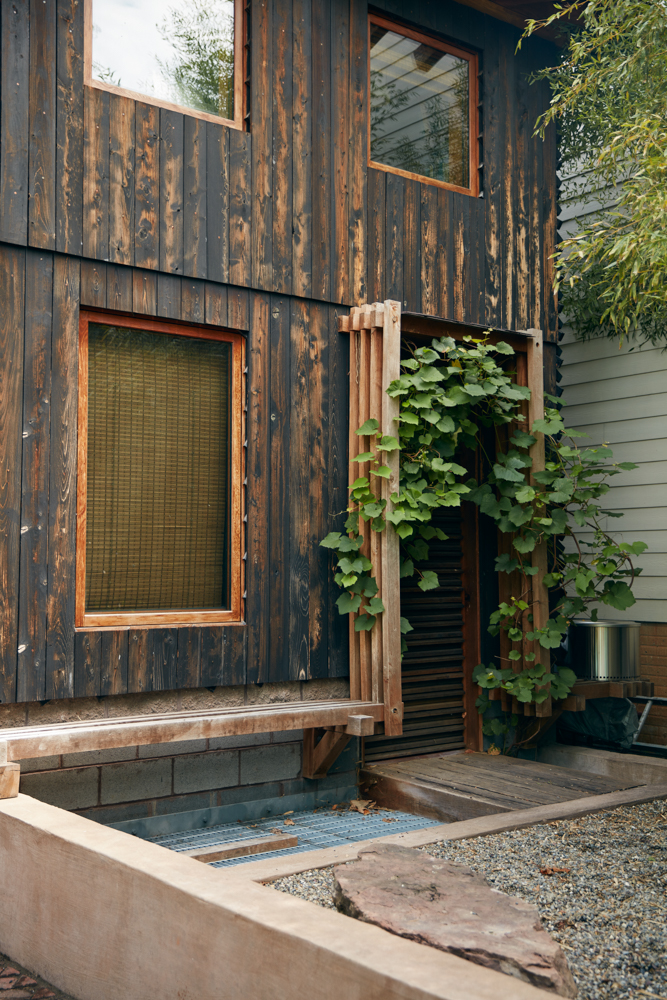 Wooden house exterior with windows, a vine-covered trellis, and a small garden area.