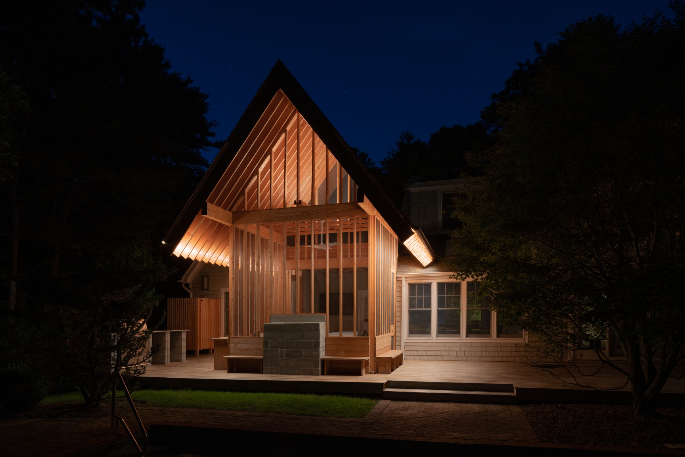 A modern house with a steep roof, illuminated at night, surrounded by trees and a deck area.