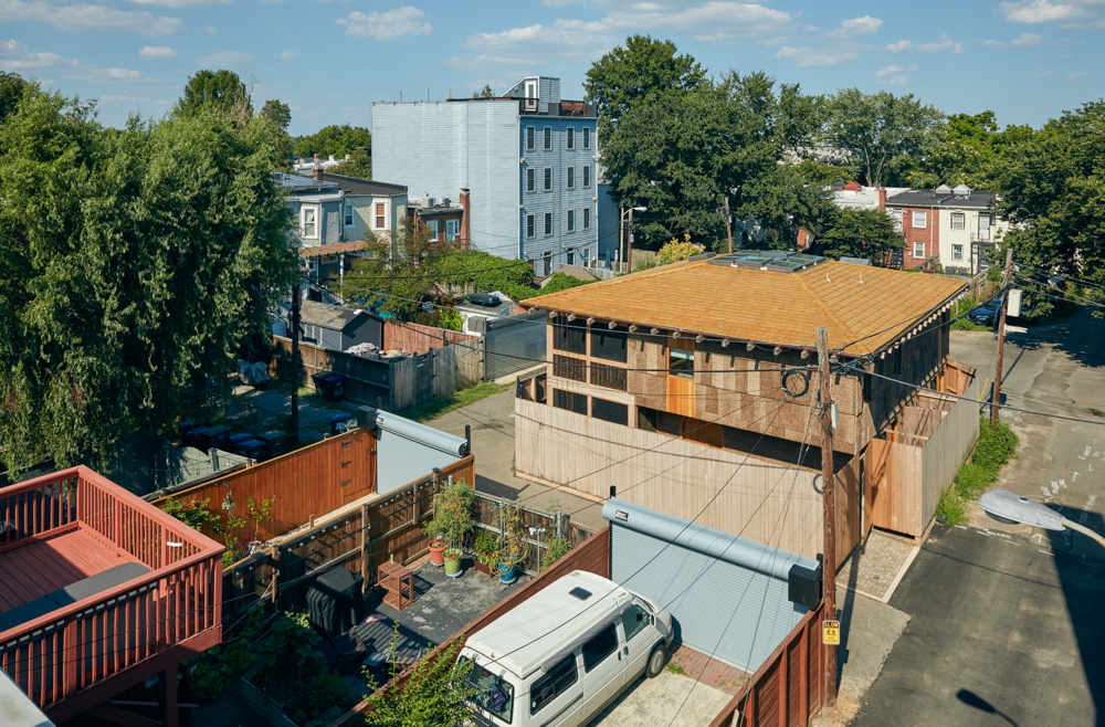 View of buildings, trees, and a van parked on a street with a wooden fence, under a blue sky with clouds.