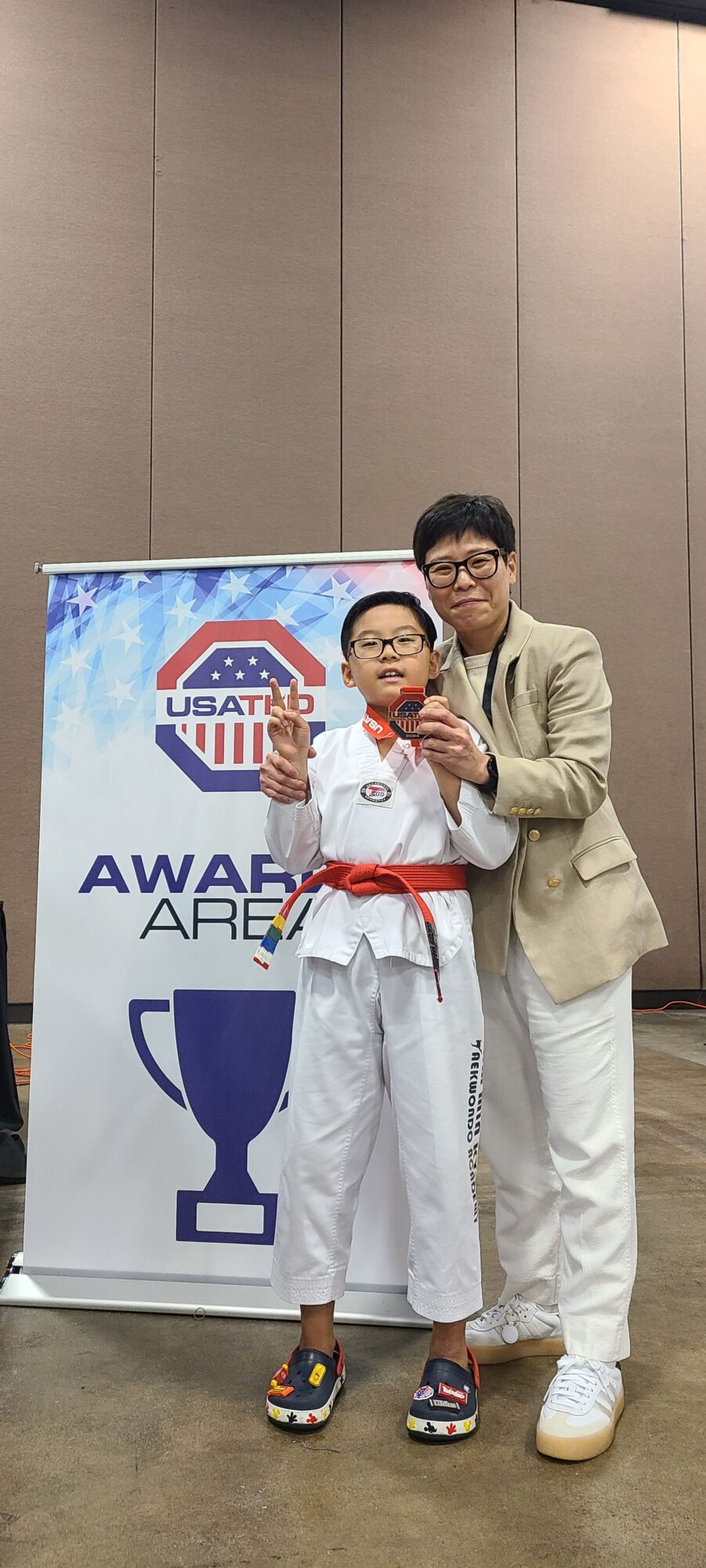 Two children in martial arts uniforms with red belts, standing with an adult in front of a sign, smiling.
