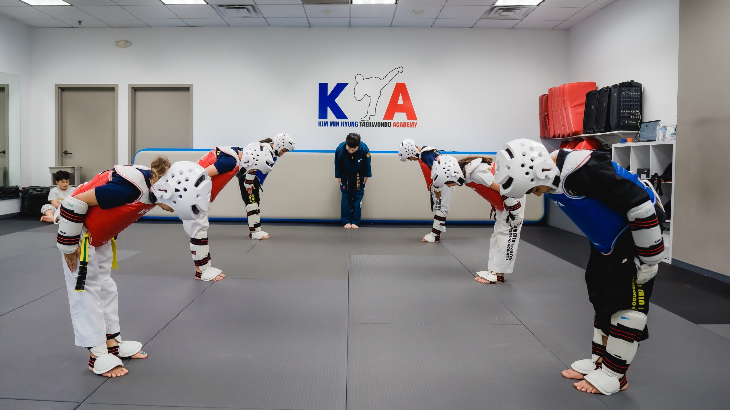 Six people in martial arts uniforms and protective gear perform a bow in a training room with a logo on the wall.