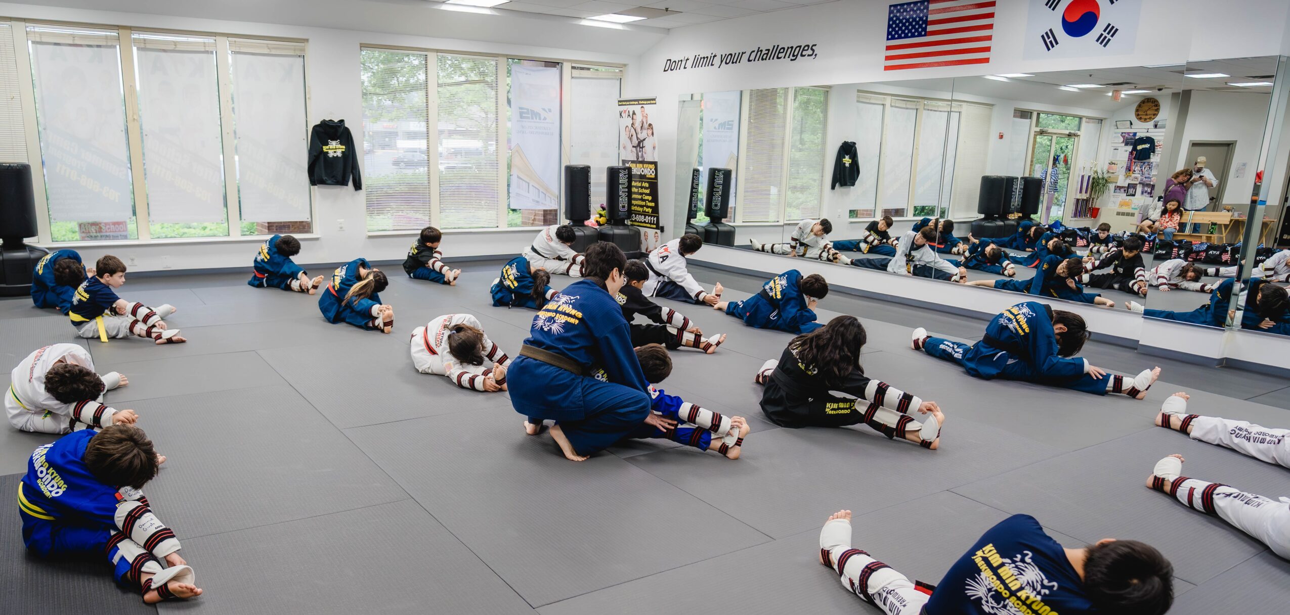 Martial arts class with children stretching on mats, instructor demonstrating, flags and mirrors in the background.
