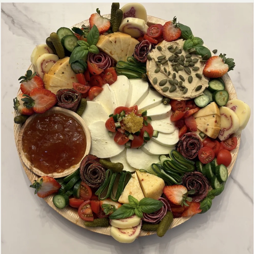 A round cake decorated with strawberries, cheese, vegetables, and edible flowers, topped with various cheeses and garnishes.