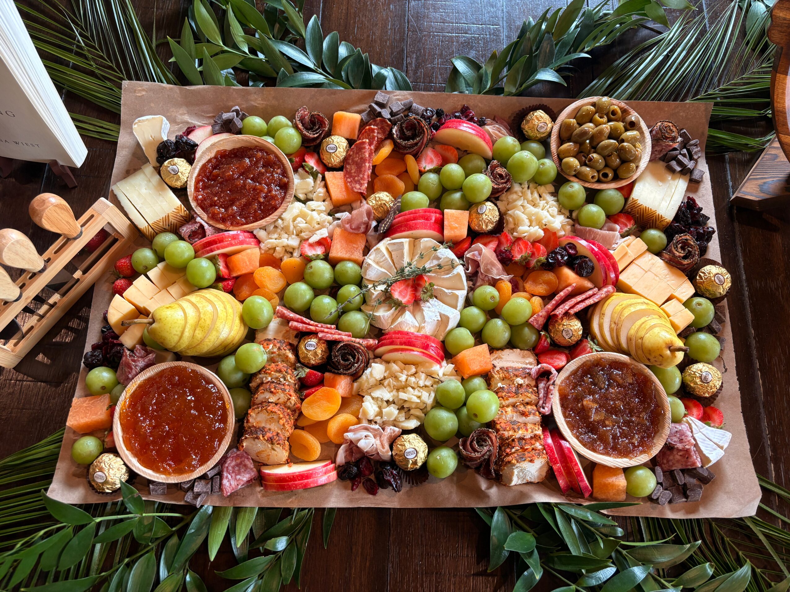 Assorted cheeses, grapes, olives, and spreads on a platter with vegetables and dried fruits, surrounded by green leaves.