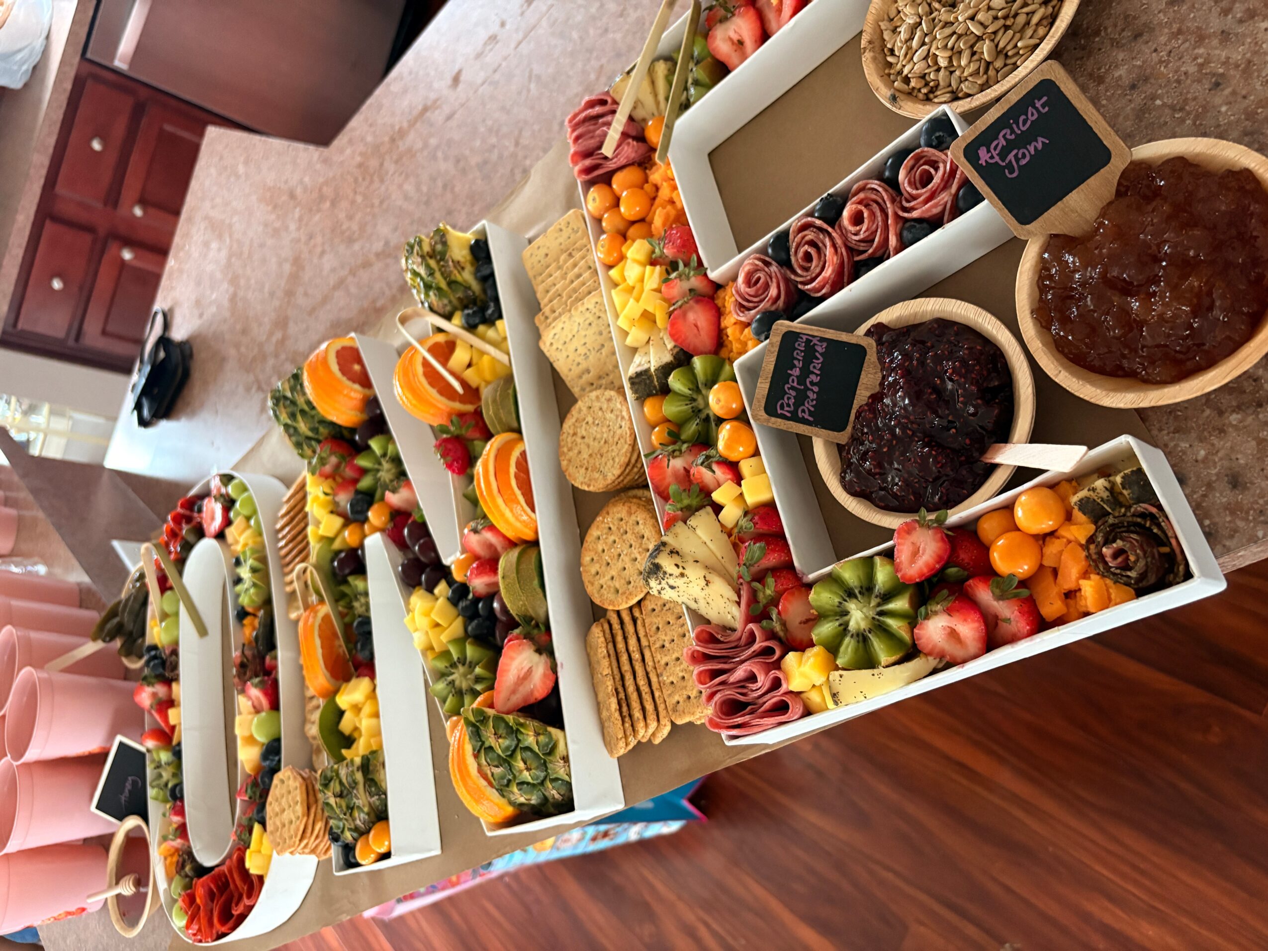 Multiple decorated boxes with fruits, nuts, and snacks on a table, with small chalkboard signs, in a kitchen setting.