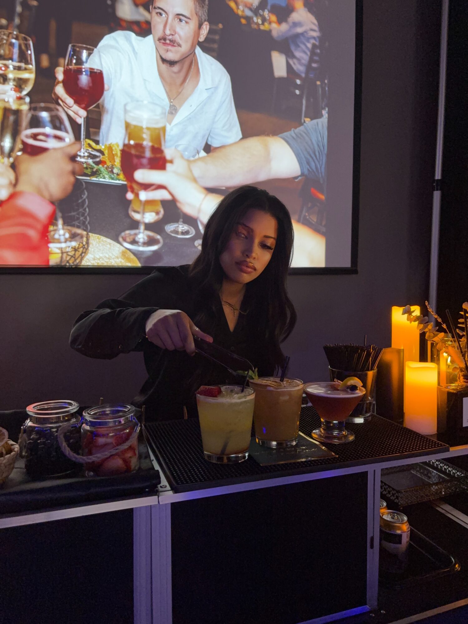 Woman preparing drinks at bar with illuminated candles and jars, large screen showing people in background, dark setting.