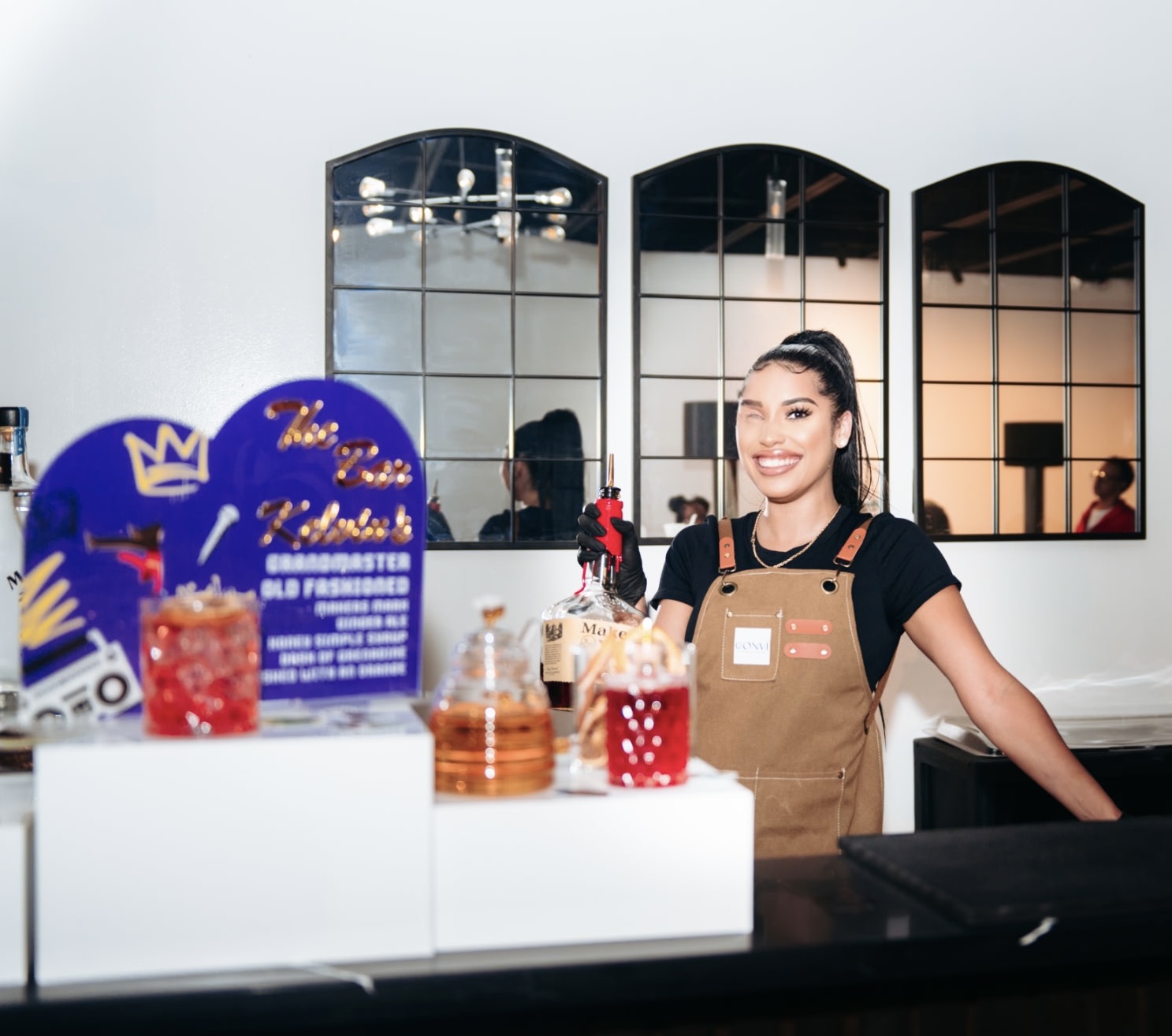 Smiling woman in apron behind a counter with drinks and decorations, mirrors in the background.