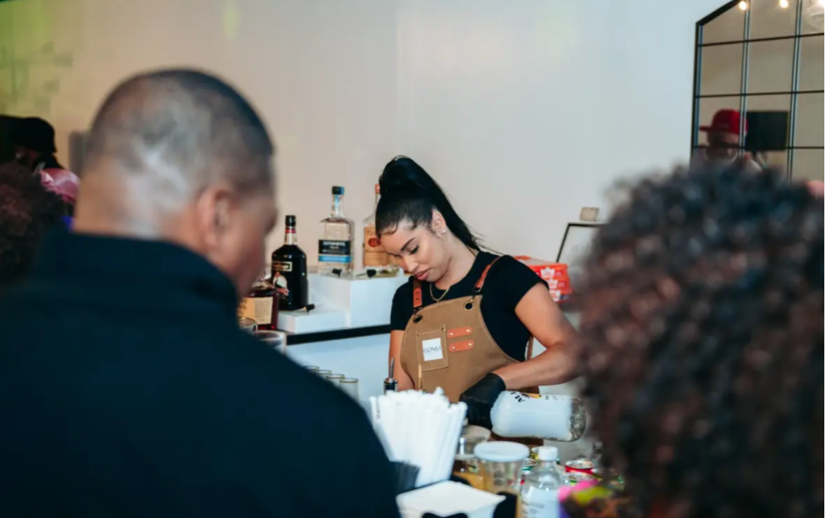 Barista preparing drinks behind counter with customers in foreground, bottles and supplies on counter, mirror on wall, neutral background.