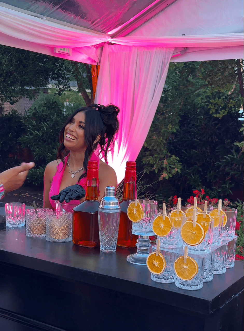 Young woman with dark hair in a bun, smiling at a party table with drinks and orange slices, under pink drapery.