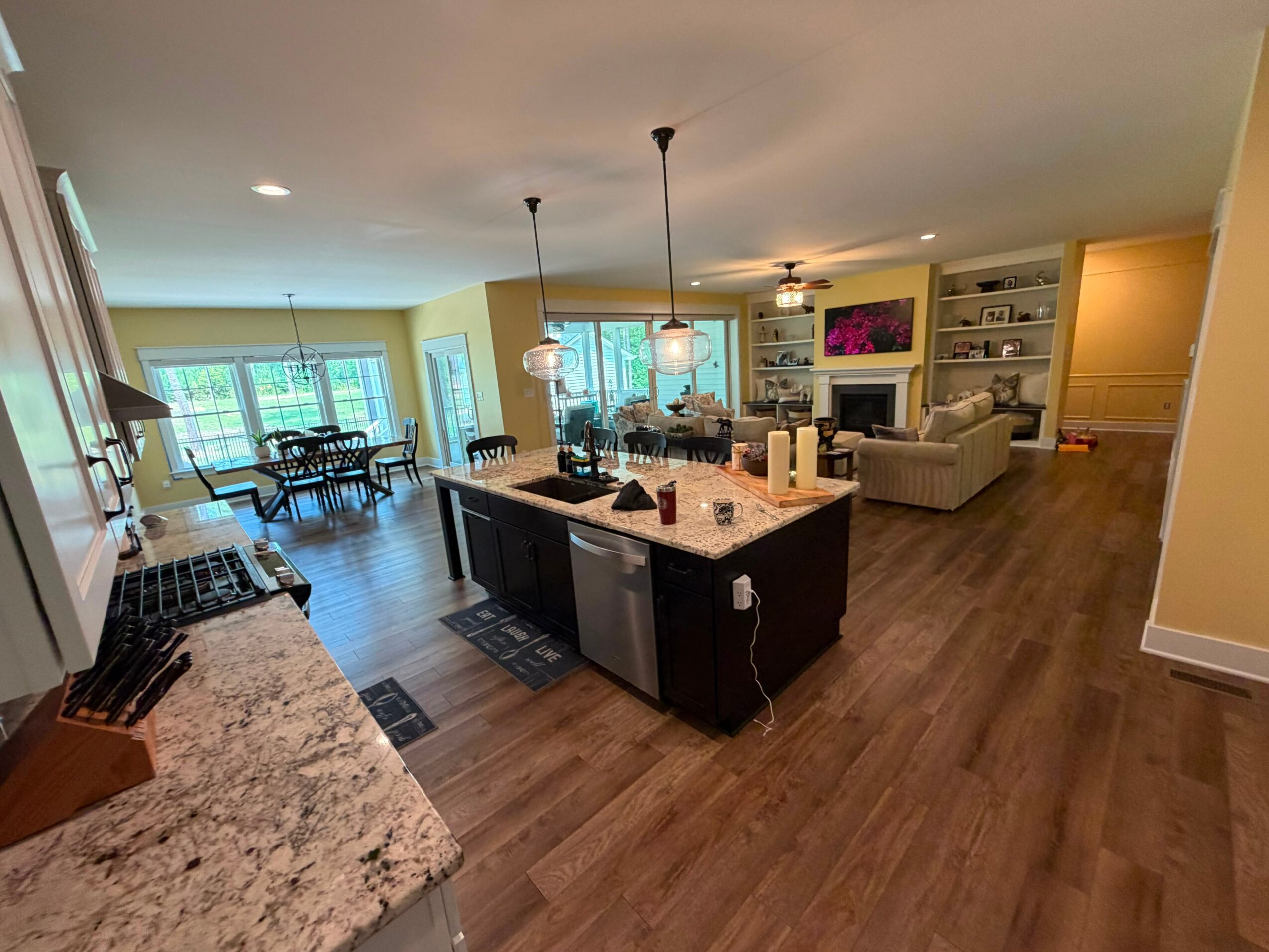 Open-concept living room and kitchen with island, dining area, and seating, illuminated by natural light and ceiling lights.