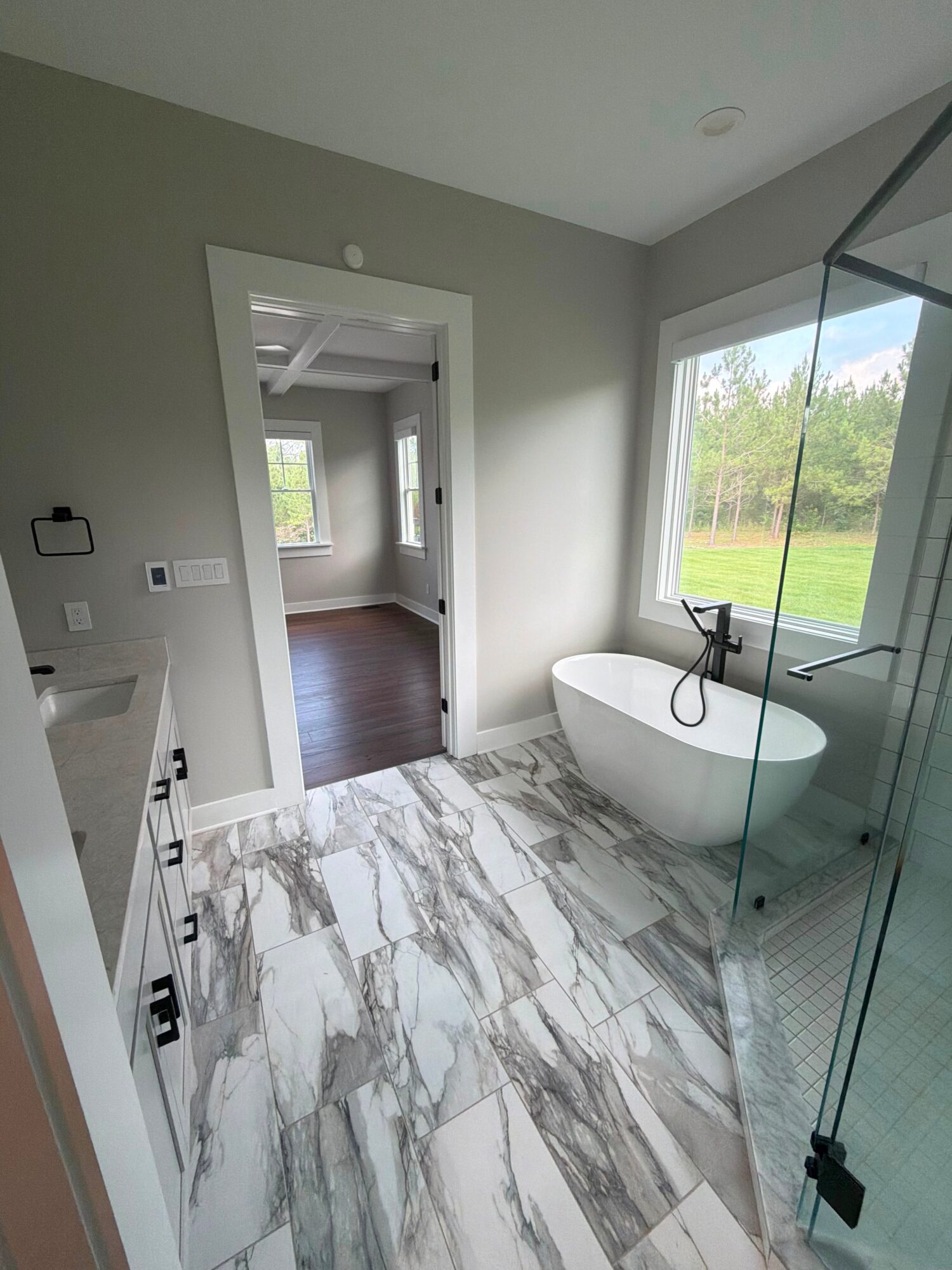 Bathroom with marble floor, freestanding bathtub near large window, glass shower enclosure, and doorway to another room.
