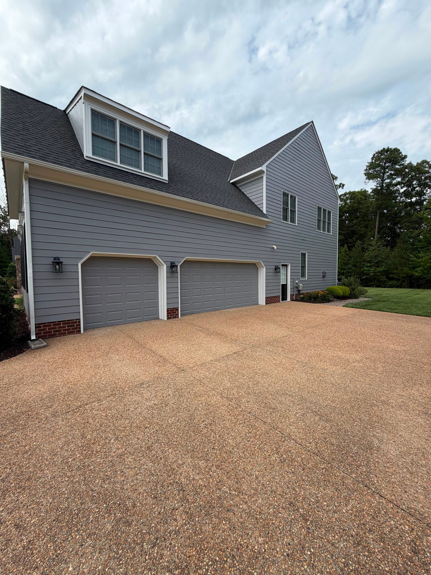 Two-story house with gray siding, two garage doors, dormer window, and a large driveway, with trees and sky in background.