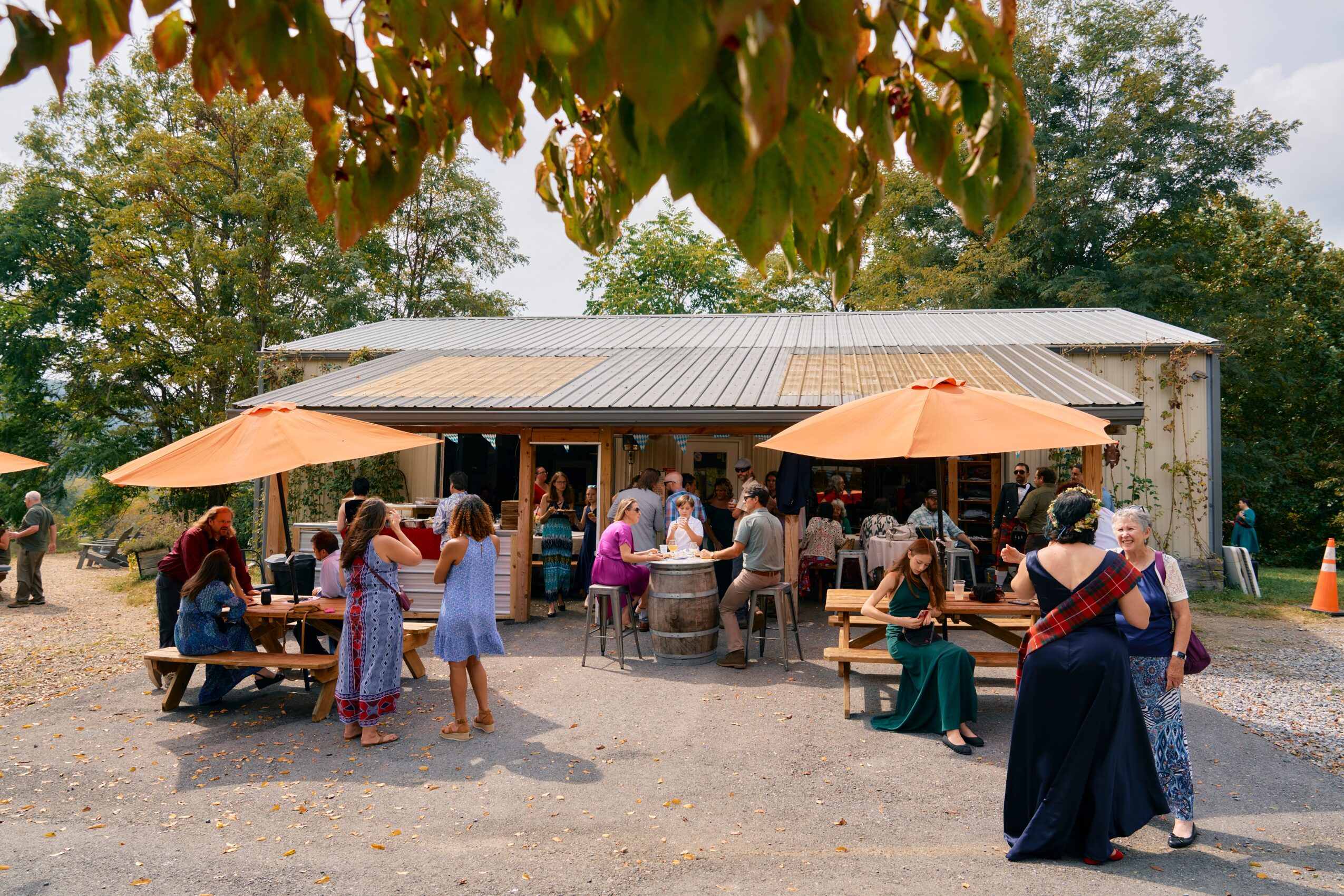 People gather outside a building with umbrellas and outdoor seating, trees in the background, and a gravel ground.