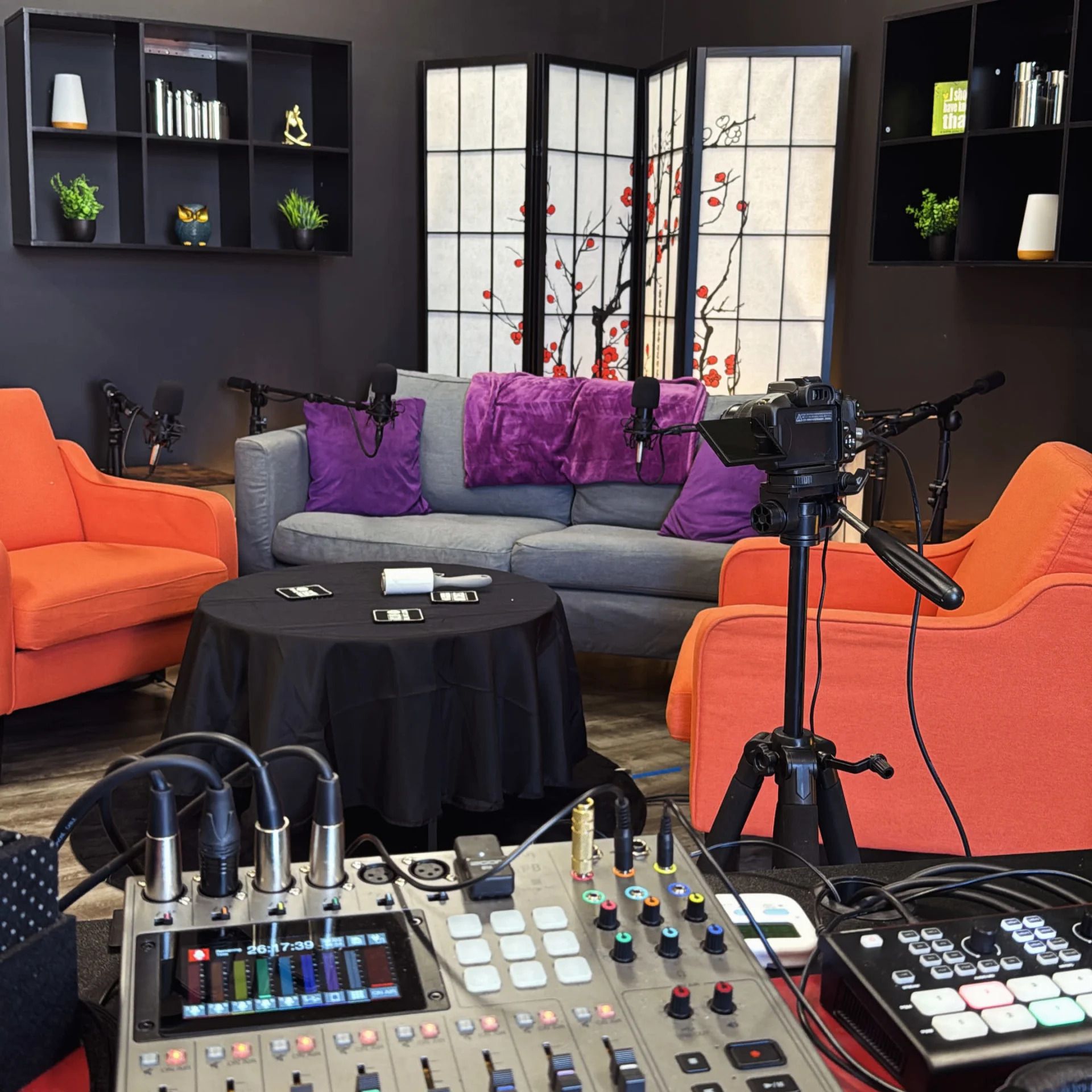 Living room with gray sofa, orange chairs, black table, camera, and audio equipment, with decorative screens and shelves in the background.