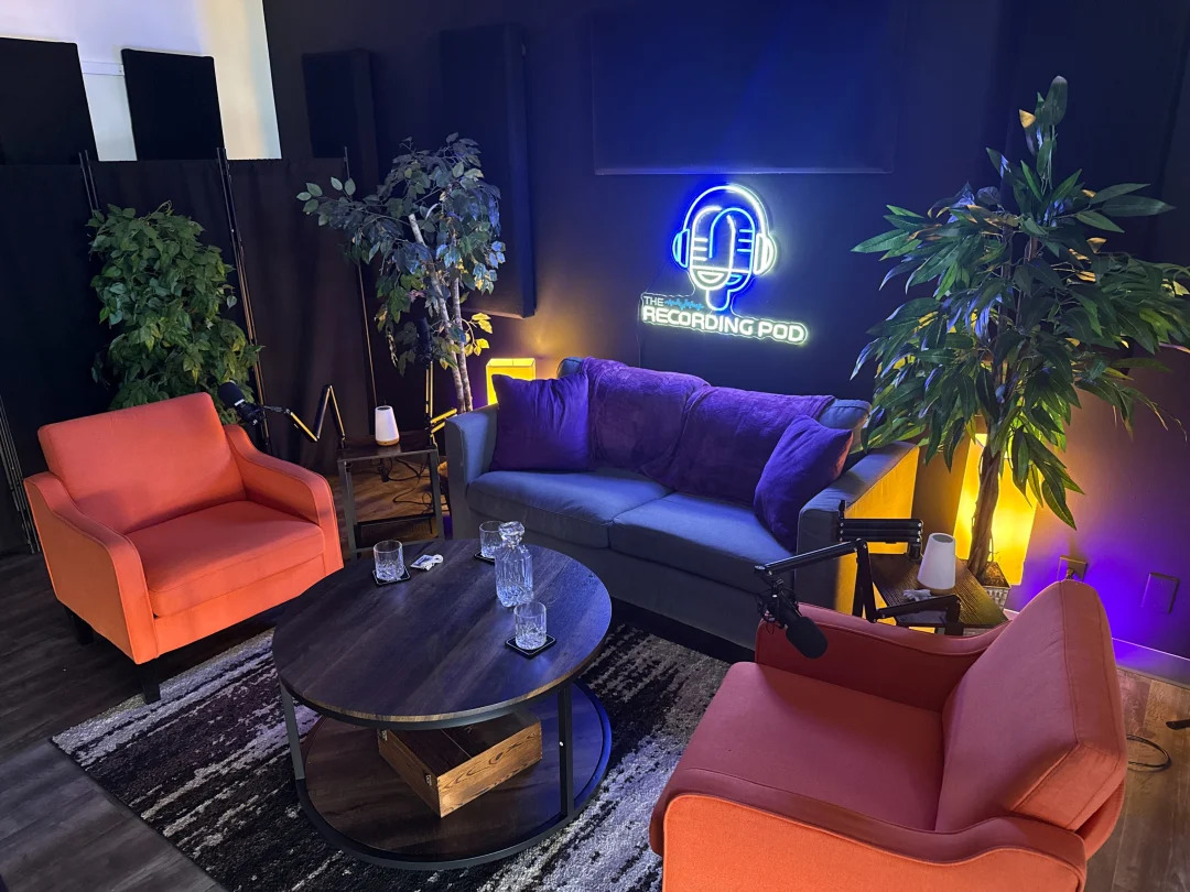 Indoor recording studio with colorful sofas, plants, a neon sign, and a black coffee table with water bottles.