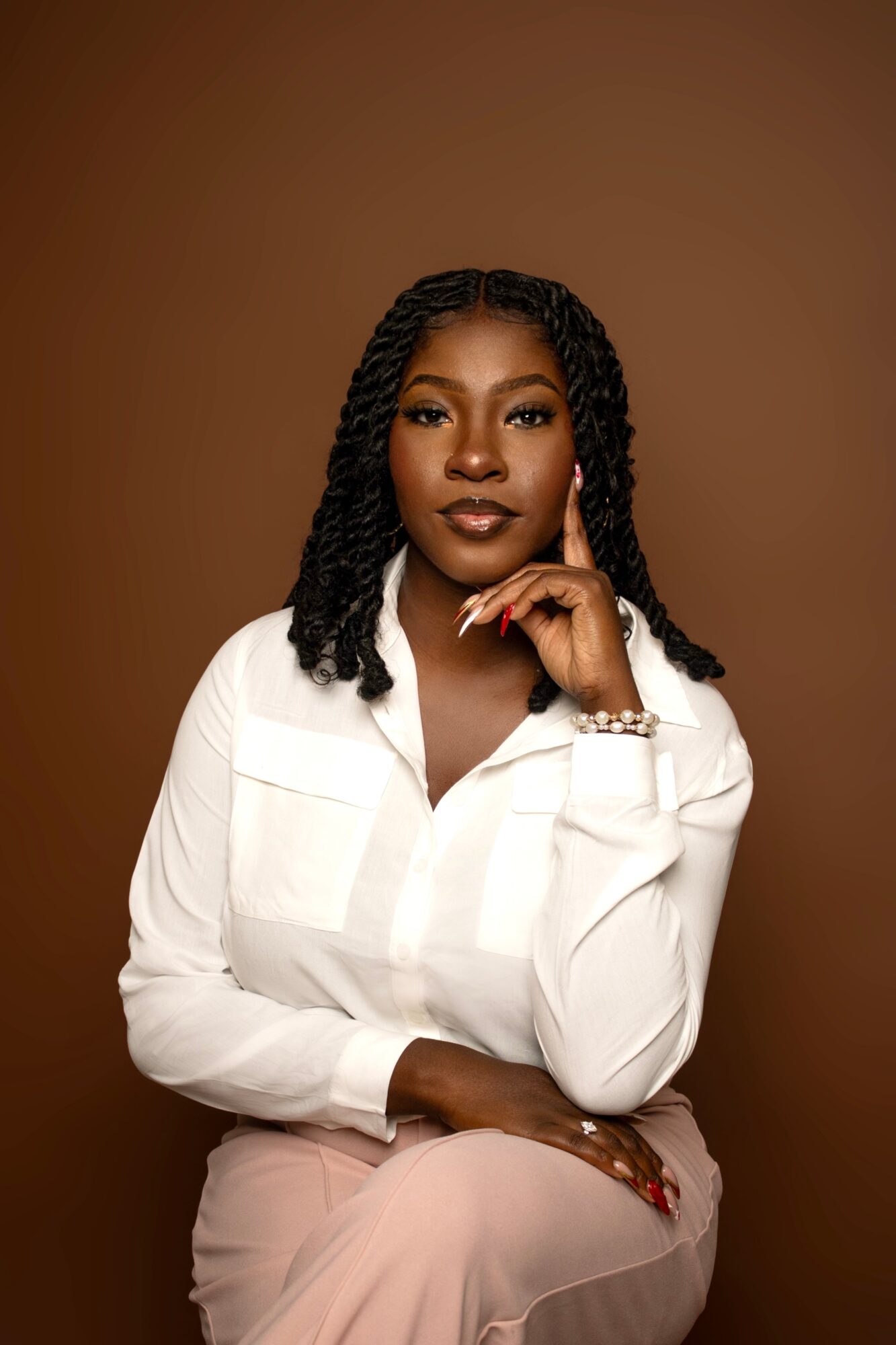 Portrait of a woman with braided hair, wearing a white shirt, sitting with her hand near her chin, against a brown background.