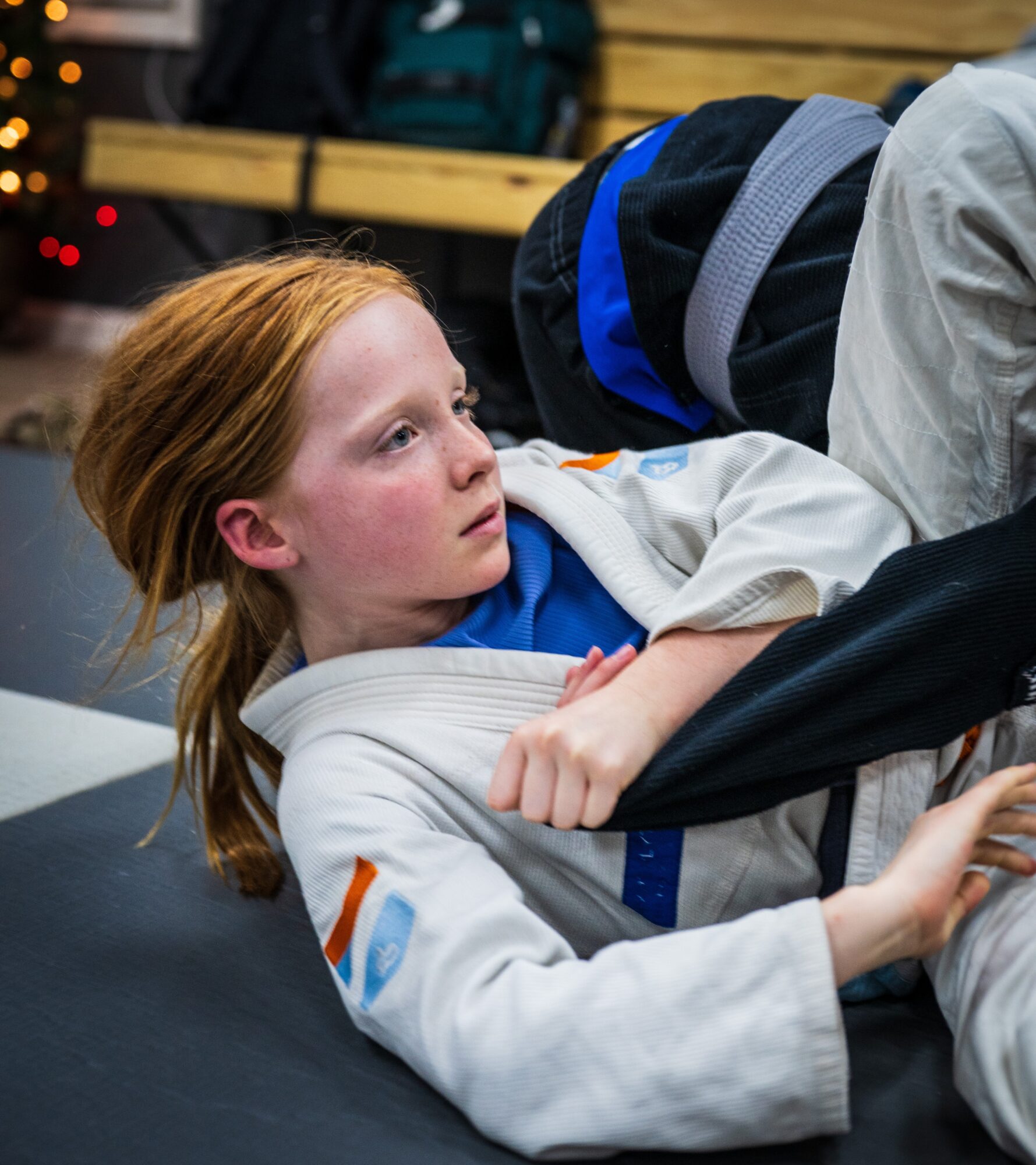 Young girl with red hair wearing a white martial arts gi, sitting on the floor indoors.