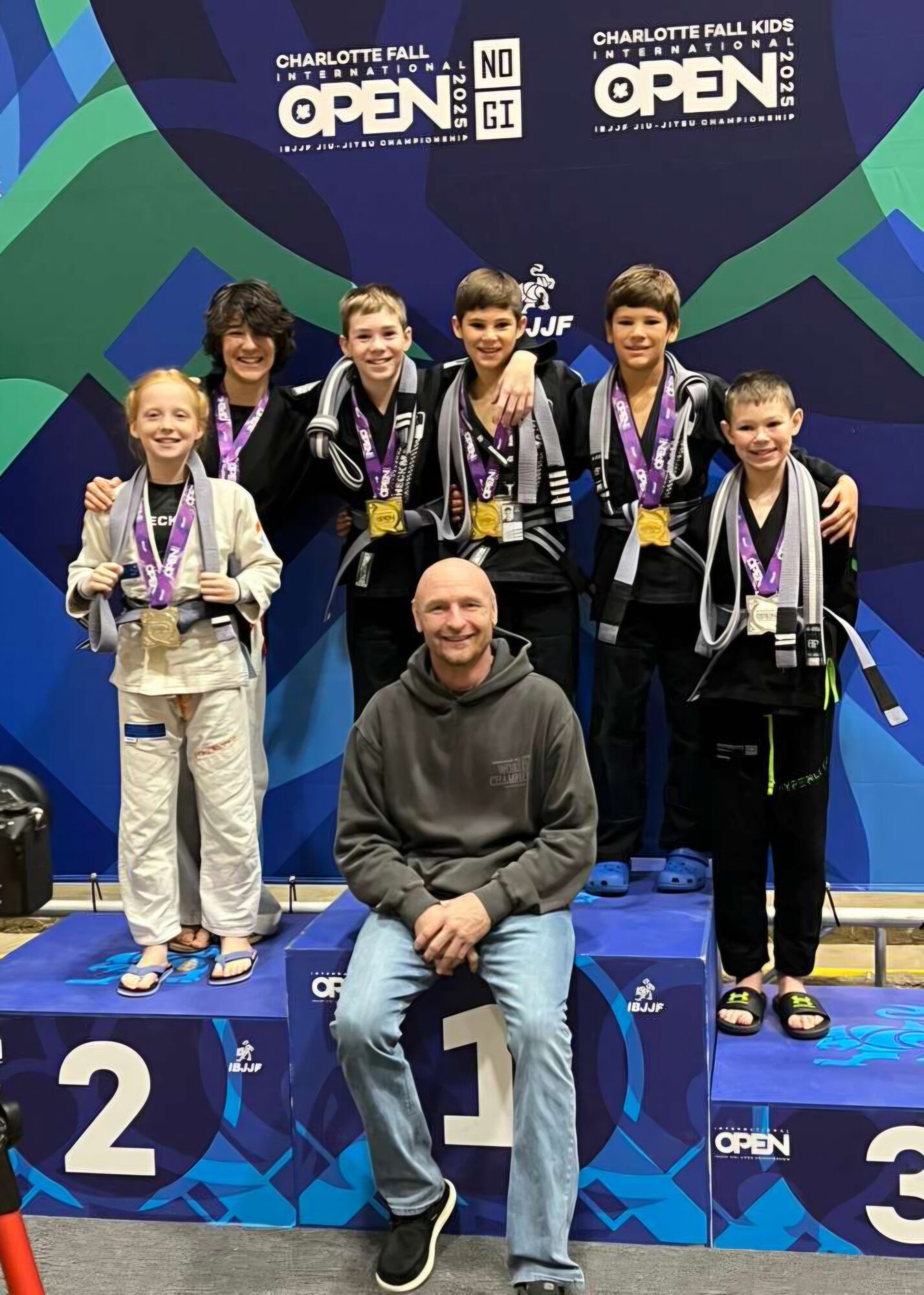 Group of children and an adult on a winners' podium with medals, smiling, in front of a colorful backdrop.