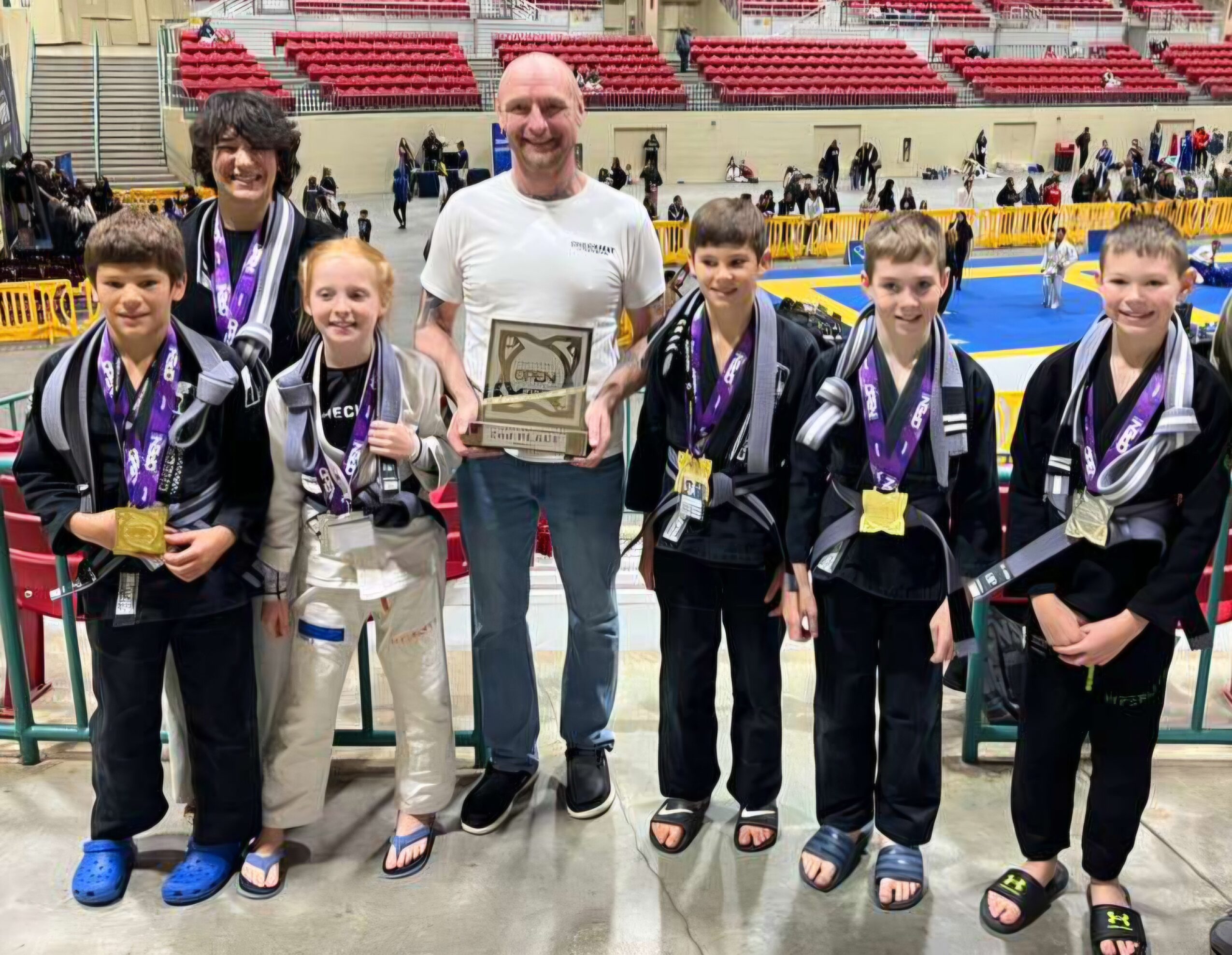 Group of six children and one adult in an indoor sports arena, holding medals and a plaque, smiling.