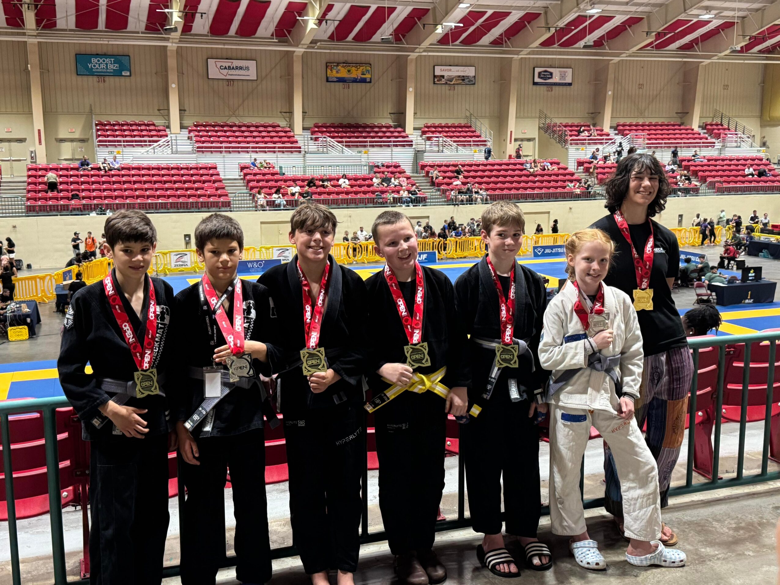 Group of six young athletes in judo uniforms with medals, standing with a coach in an indoor sports arena.