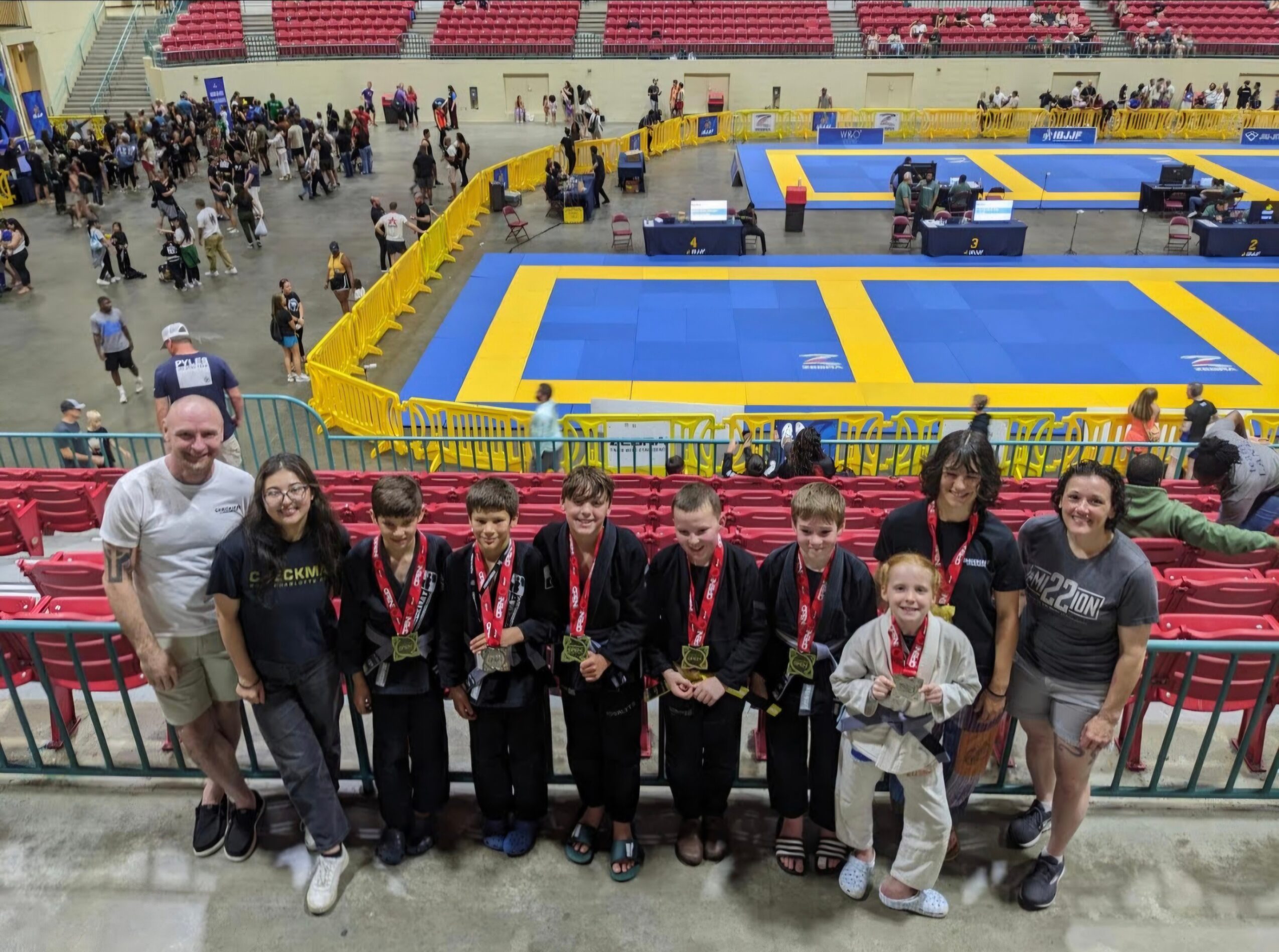 Group of young athletes with coaches in front of a martial arts competition area, some holding medals, in an indoor arena.