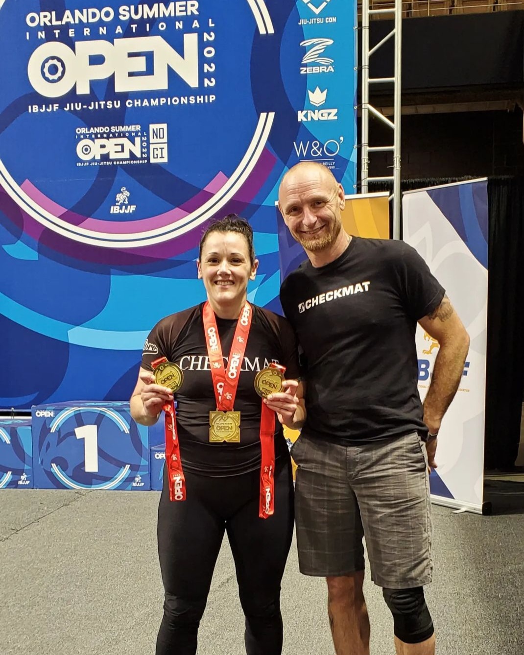 Two people standing on a stage with a large Orlando Summer Open banner behind them, smiling and holding medals.