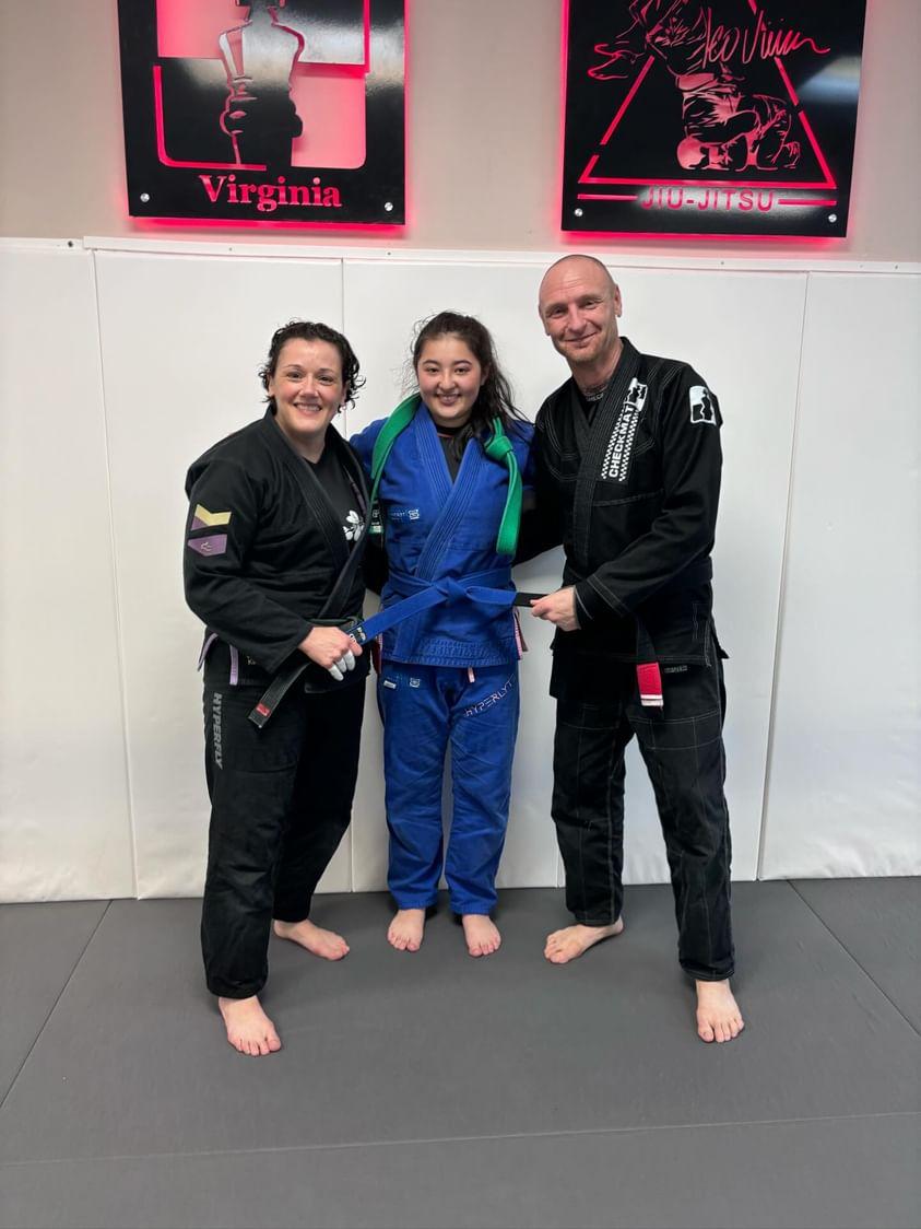 Three people in Brazilian Jiu-Jitsu gis standing on a mat, smiling, with posters on the wall behind them.
