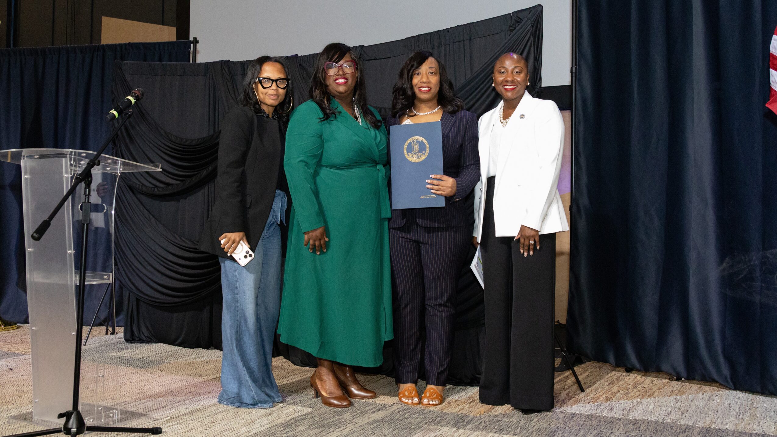 Four women standing on stage, one holding a certificate, with a black curtain backdrop and a microphone stand nearby.