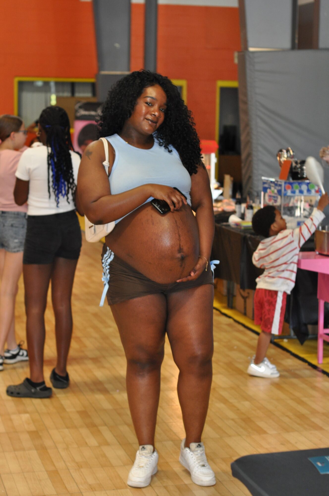 Person with dark skin and curly hair standing in a room with other people, some wearing casual clothing, wooden floor, colorful walls.