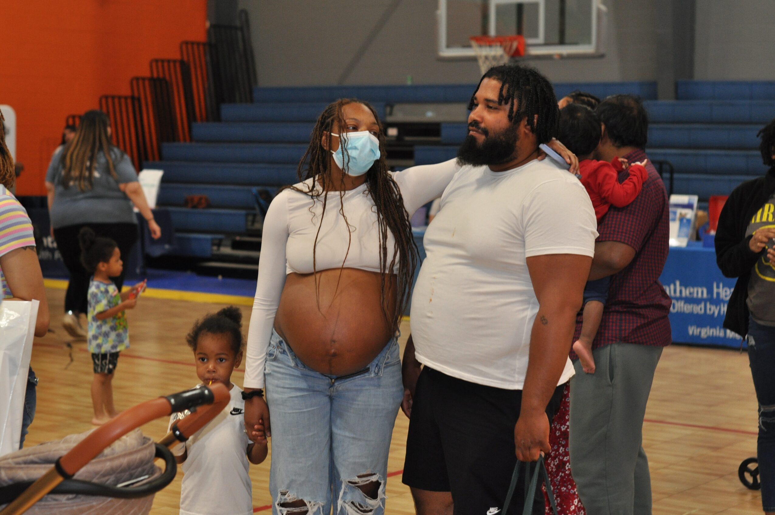 Pregnant woman and man standing together in a gymnasium, woman wearing a face mask, others in the background.
