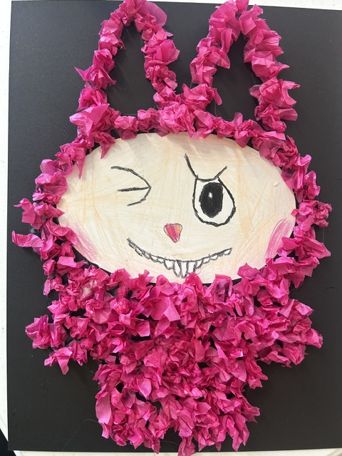 A handmade mask with a white face, black eye, pink nose, and a stitched mouth, surrounded by pink crumpled paper.