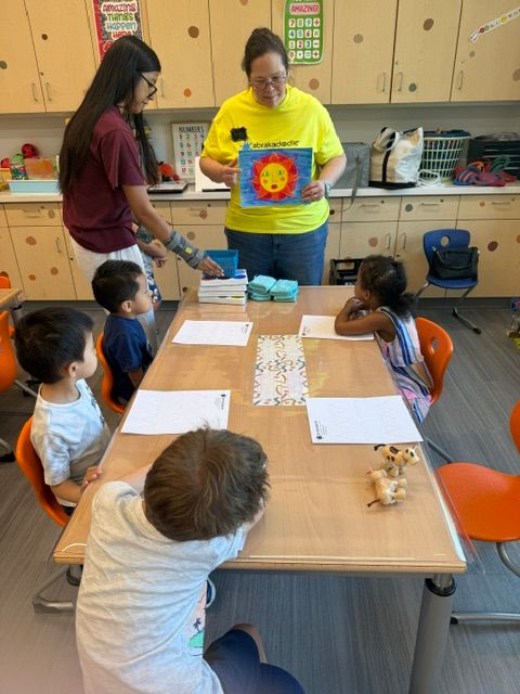 Group of children and an adult sitting around a table with papers and a toy, in a classroom setting.