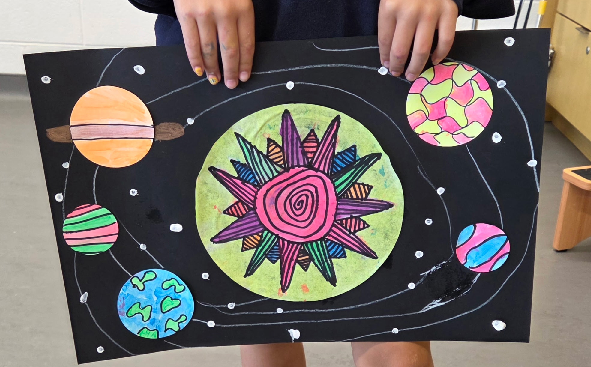 Child holds a colorful space-themed drawing with planets, stars, and orbit lines on black background.