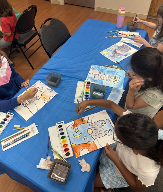 Children sit around a blue table painting colorful pictures with watercolors and brushes, with art supplies scattered on the table.