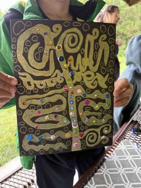 Person holding a black and gold abstract artwork with colorful dots, outdoors with grass and a person in the background.