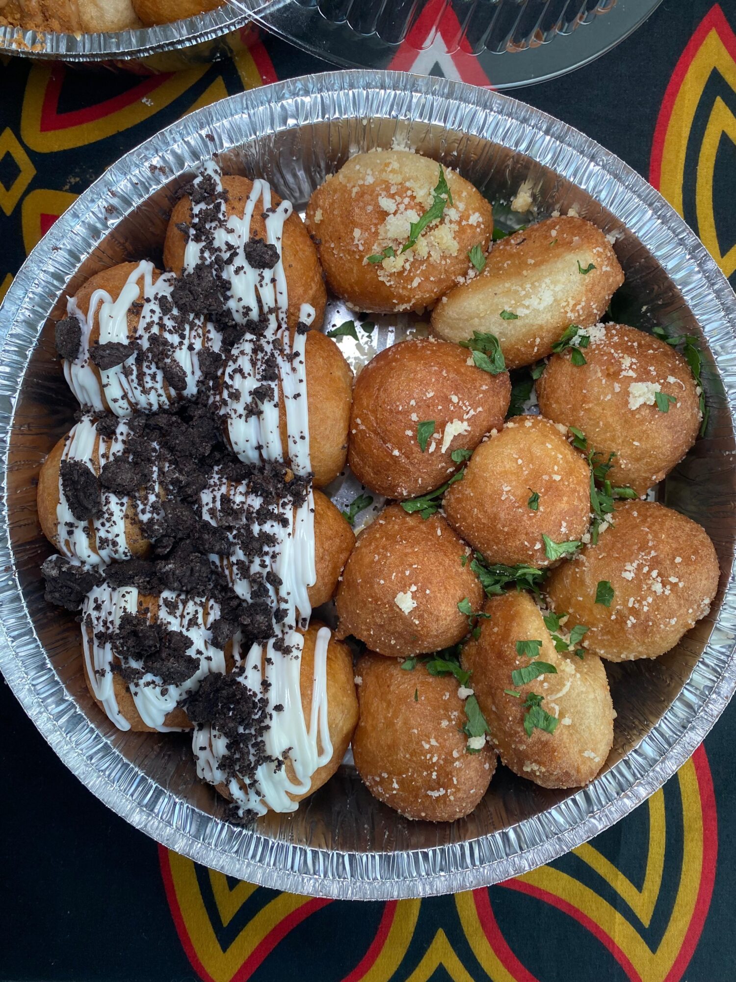 Fried snacks topped with chopped green herbs, served with a side of cookies drizzled with white and chocolate toppings in a round container.