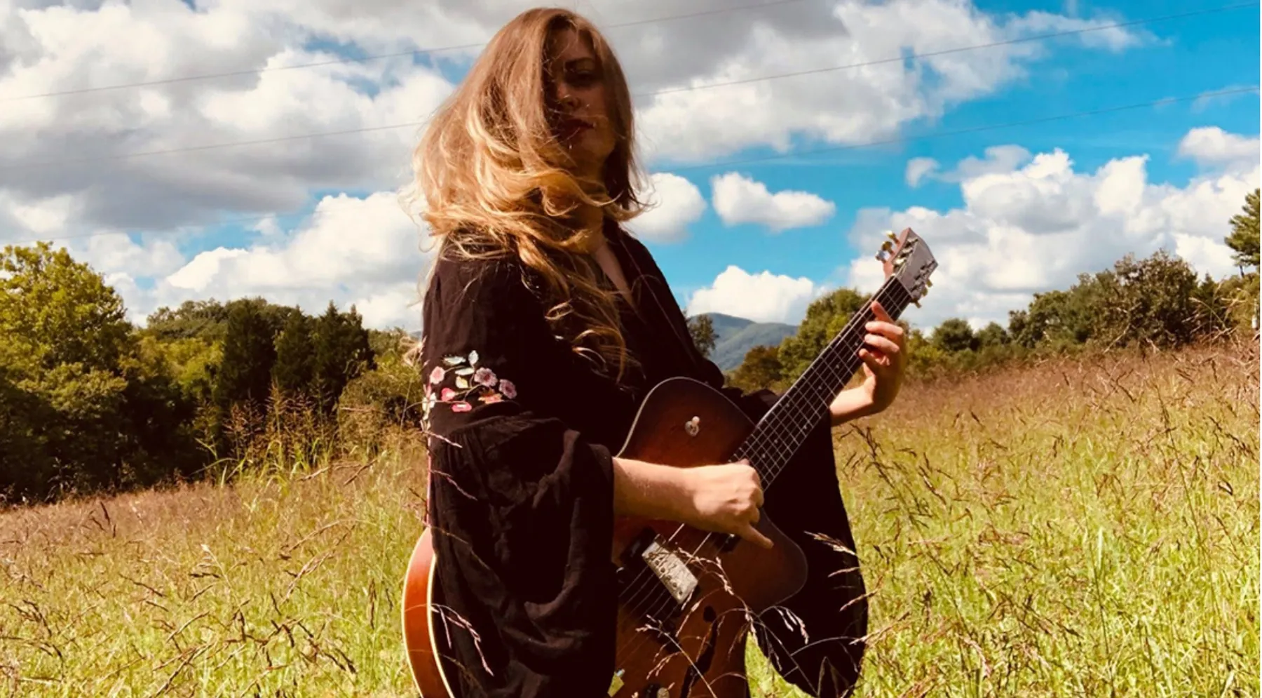Person with long hair holding a guitar in a grassy field under a partly cloudy sky